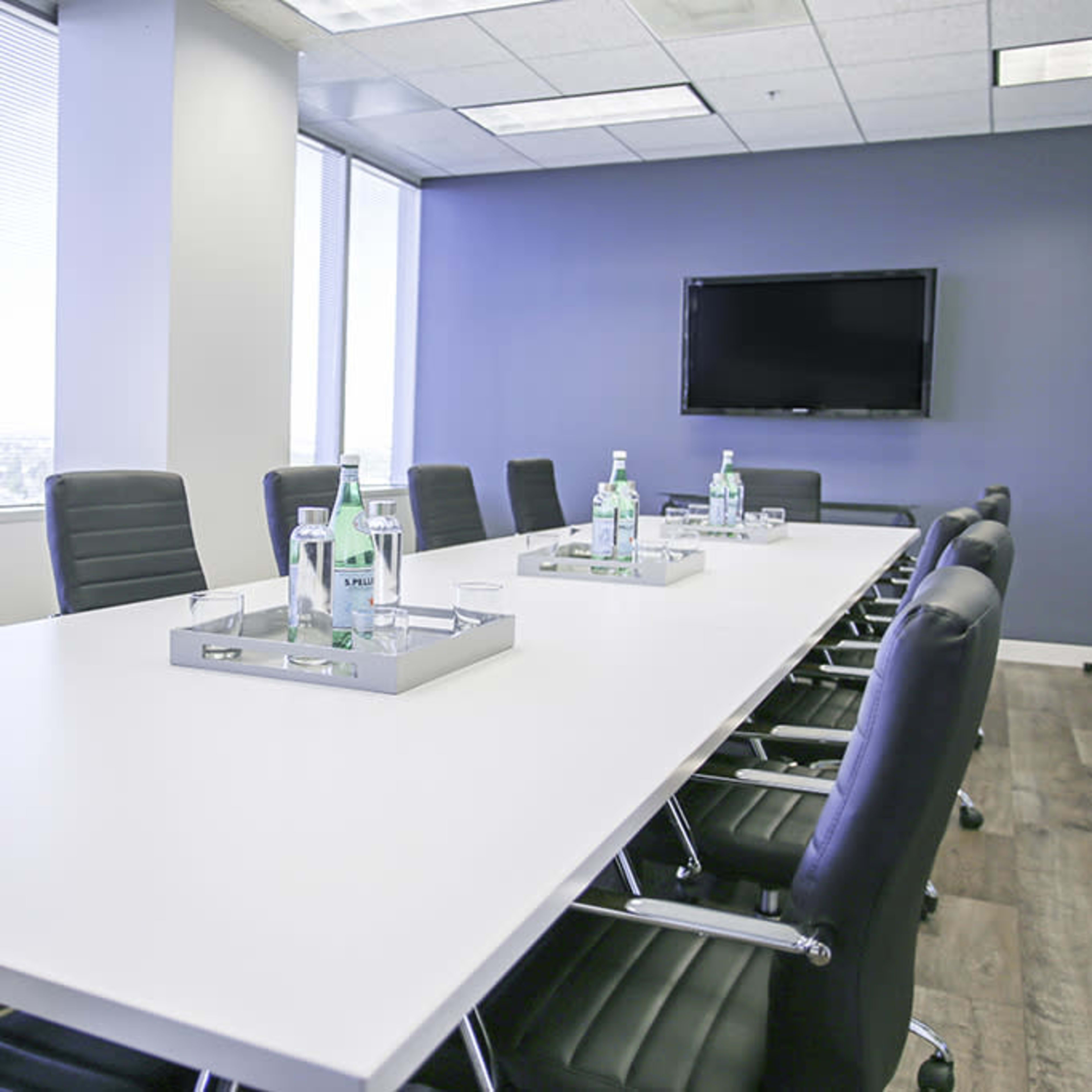 A conference room features a long white table with black chairs, surrounded by windows and a television mounted on a blue wall.