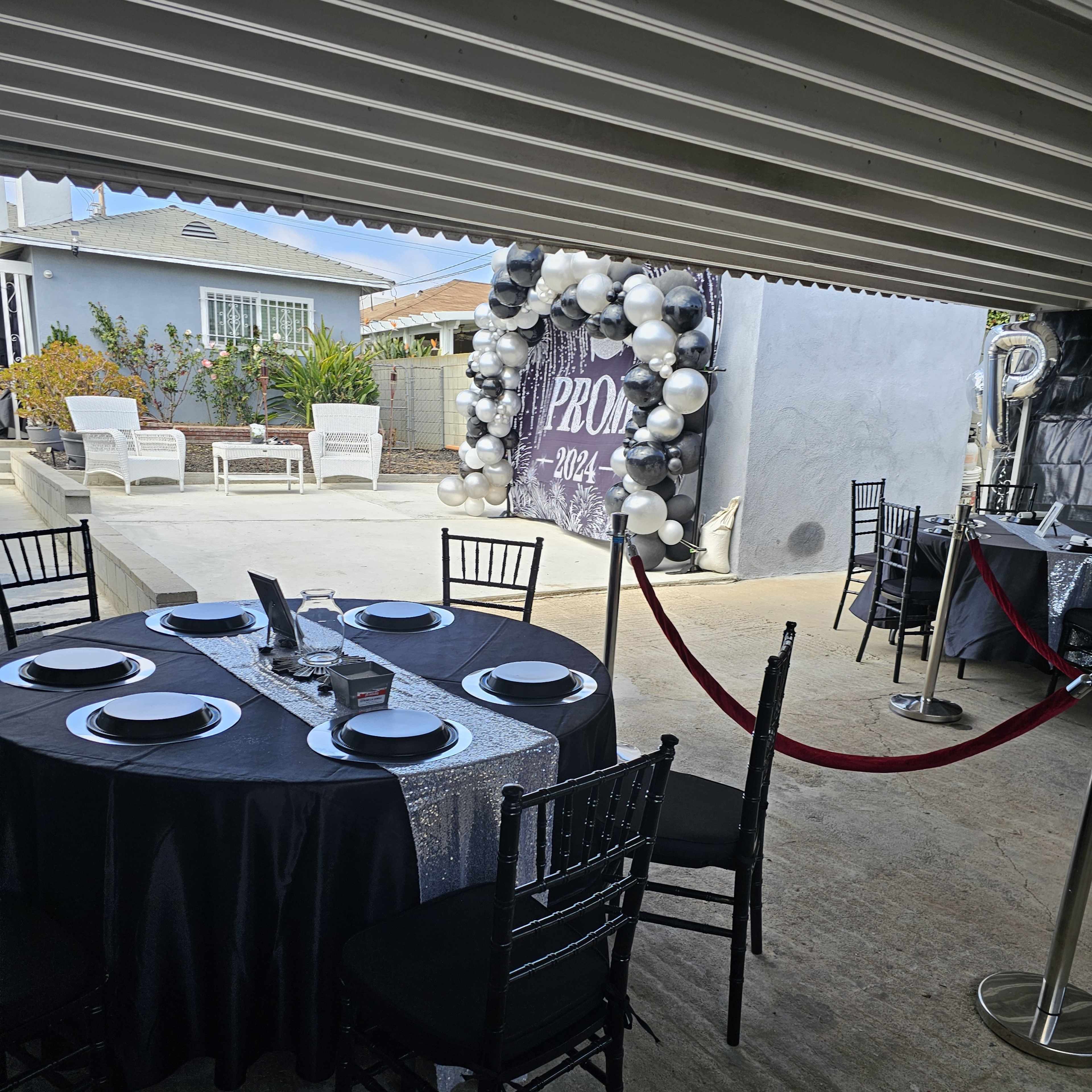 A round black table is set for an event, surrounded by black chairs, with a decorative archway of balloons in the background.
