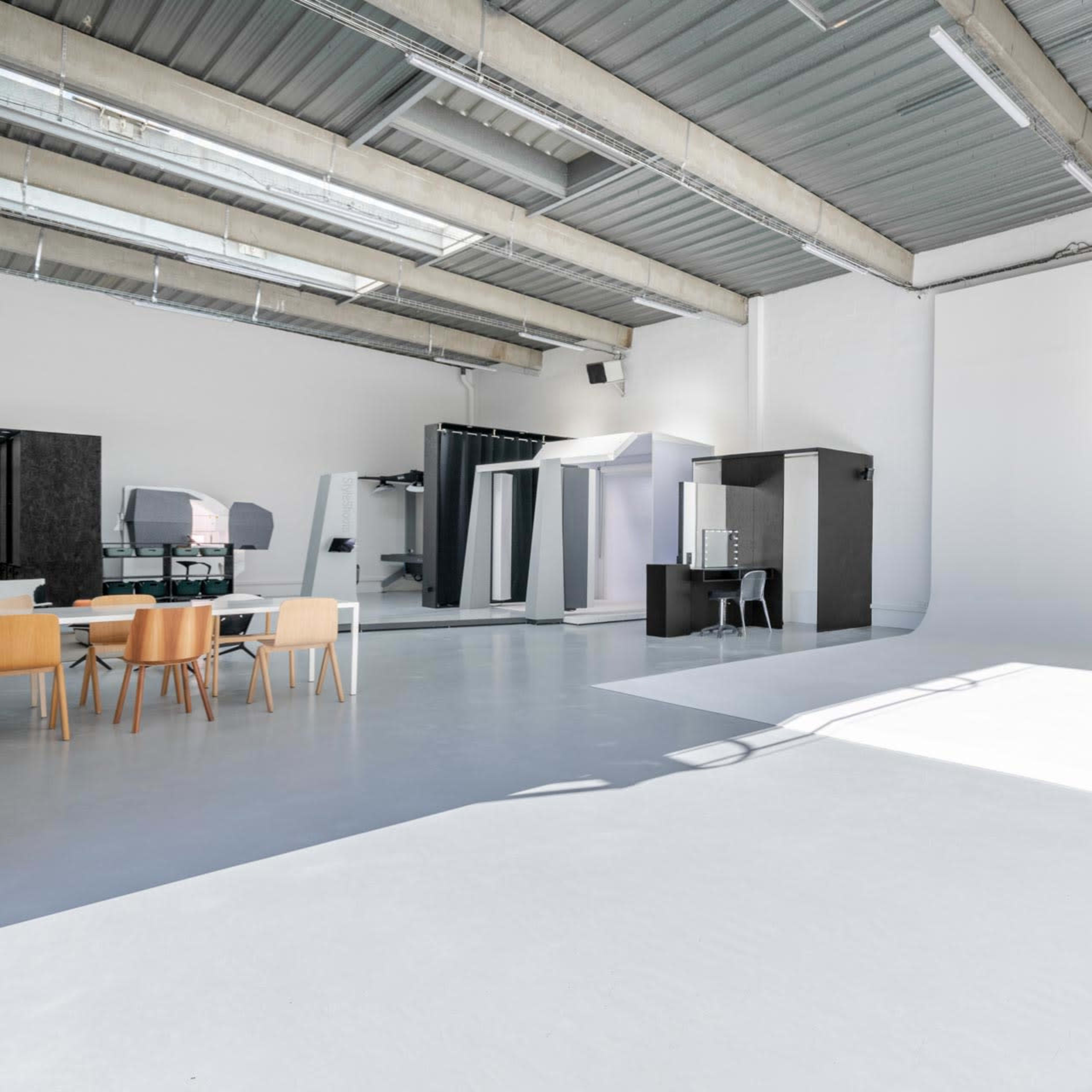 A bright, empty studio space featuring a white backdrop, a dining table with four chairs, and various furnishings against the walls.