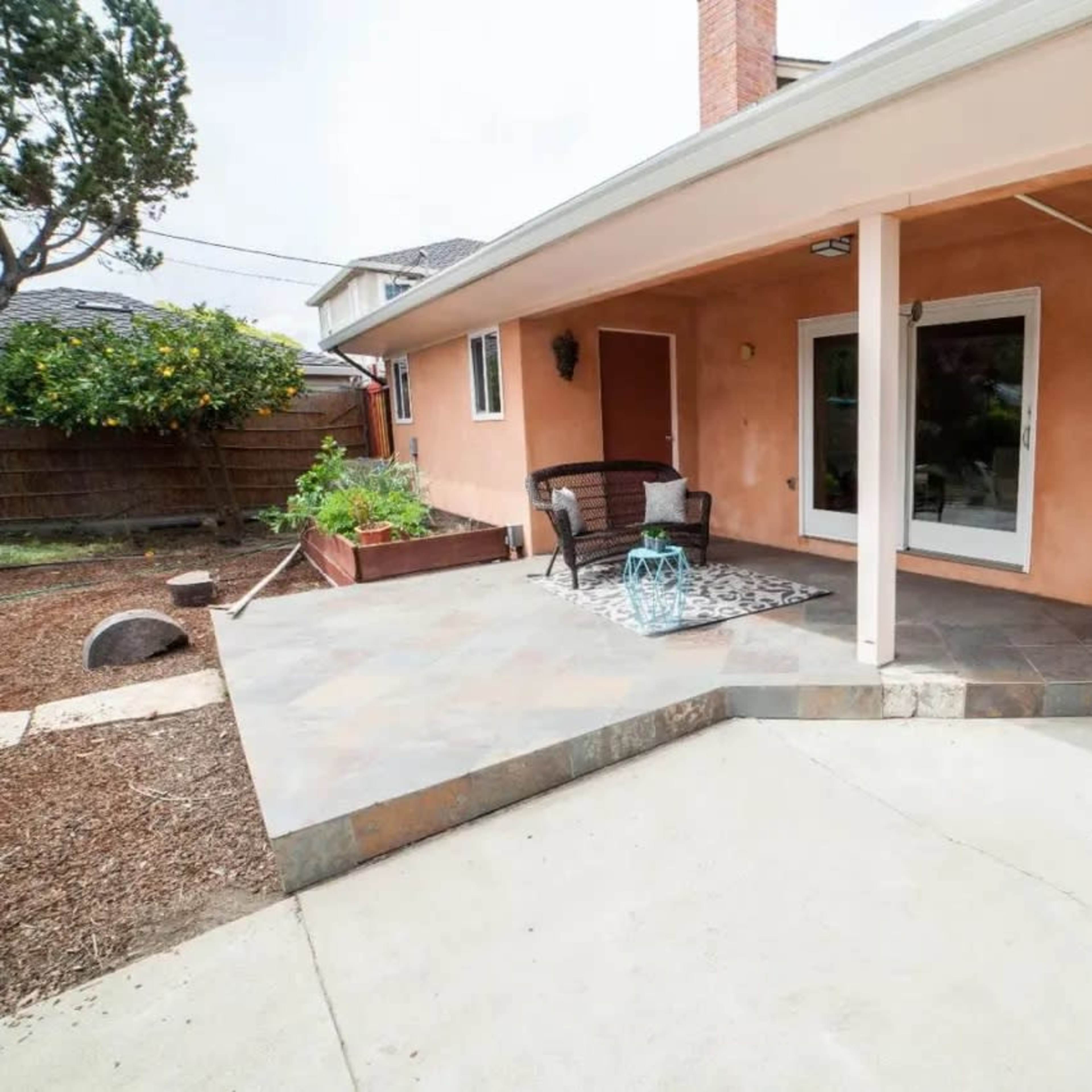 A patio area of a house featuring a tiled surface, a small table with chairs, and a landscaped yard with a wooden fence.