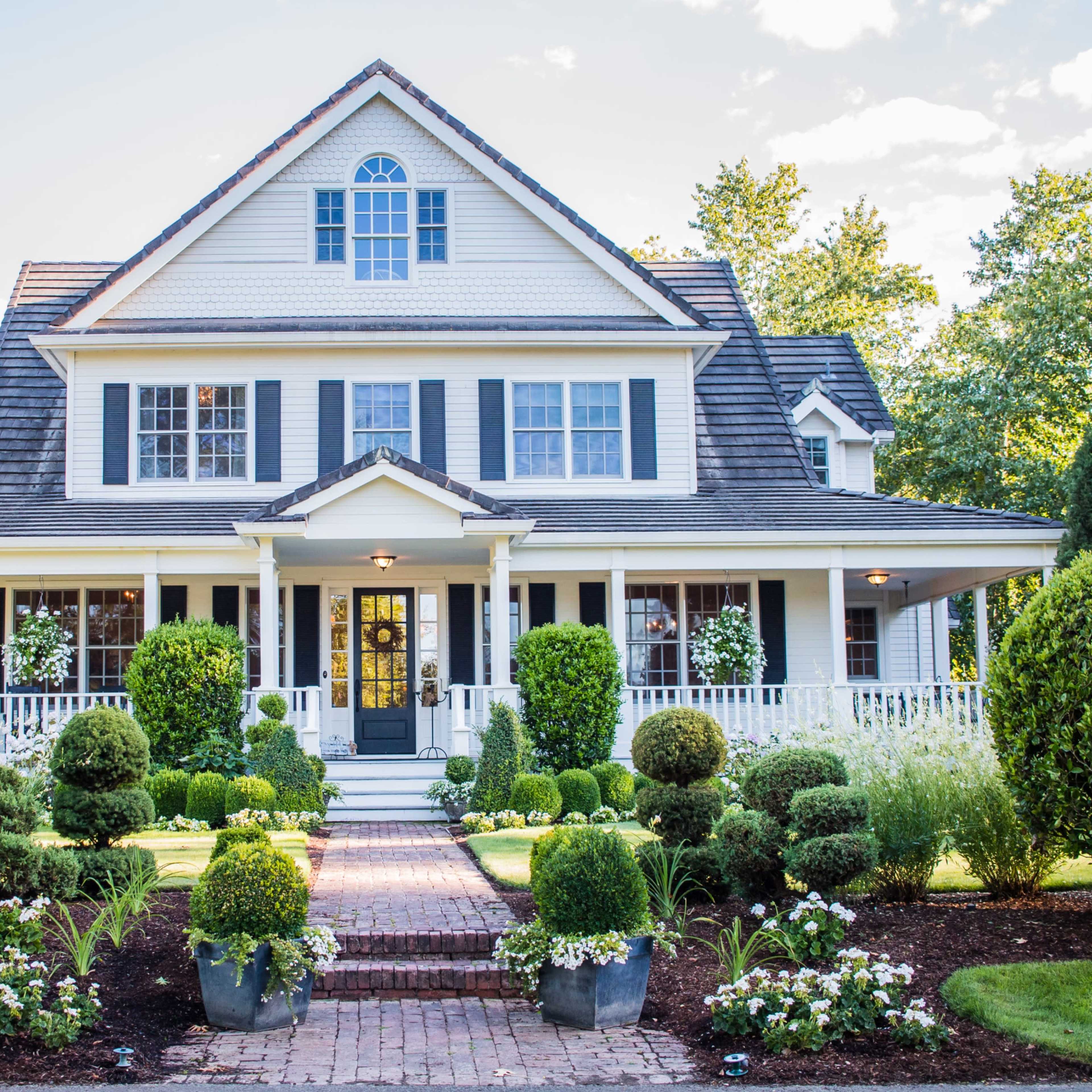 A large, two-story house with a steeply pitched roof and a front porch is surrounded by well-maintained shrubs and flowerbeds.