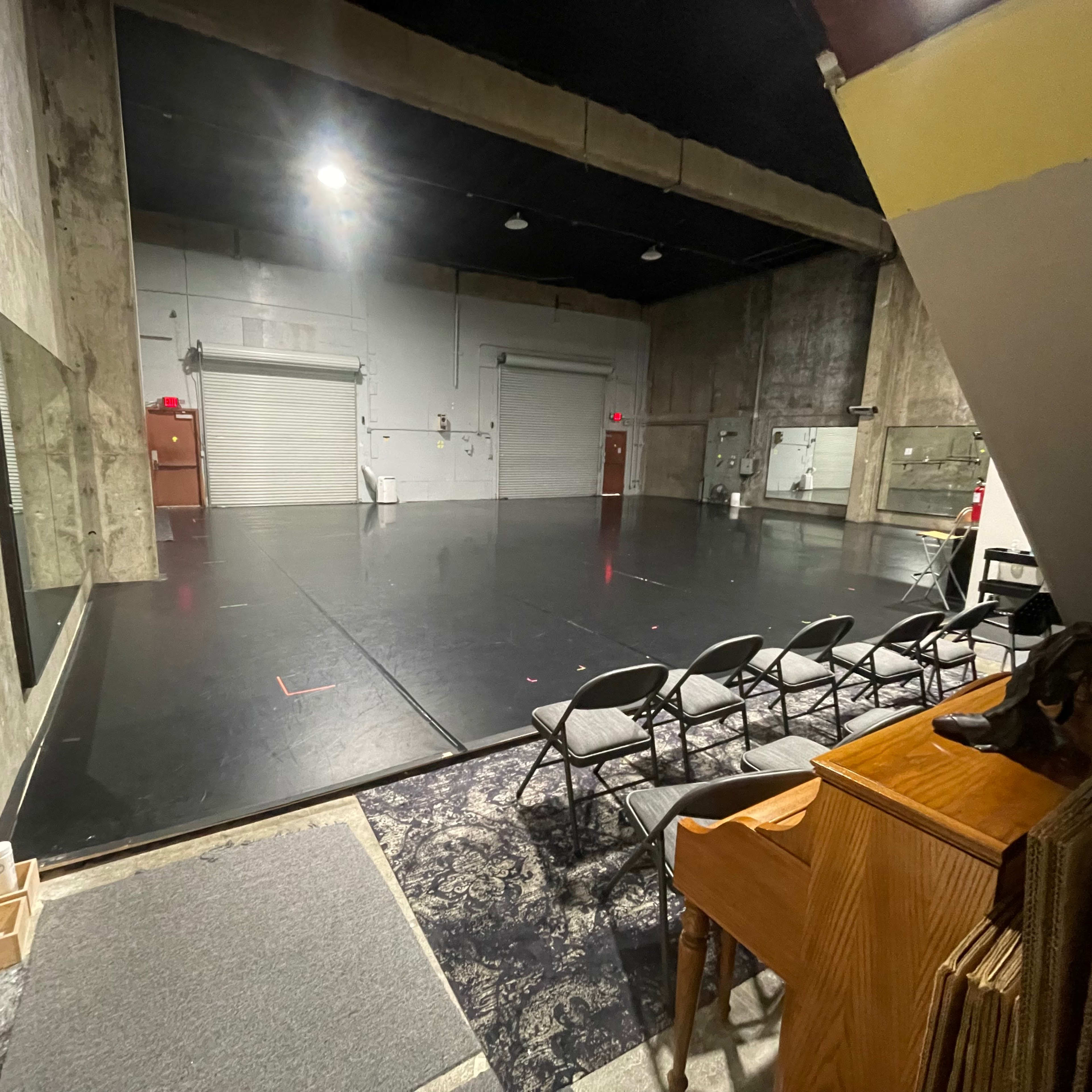 An empty rehearsal studio features a mirrored wall, a black floor, and folding chairs arranged in front of a small stage area.