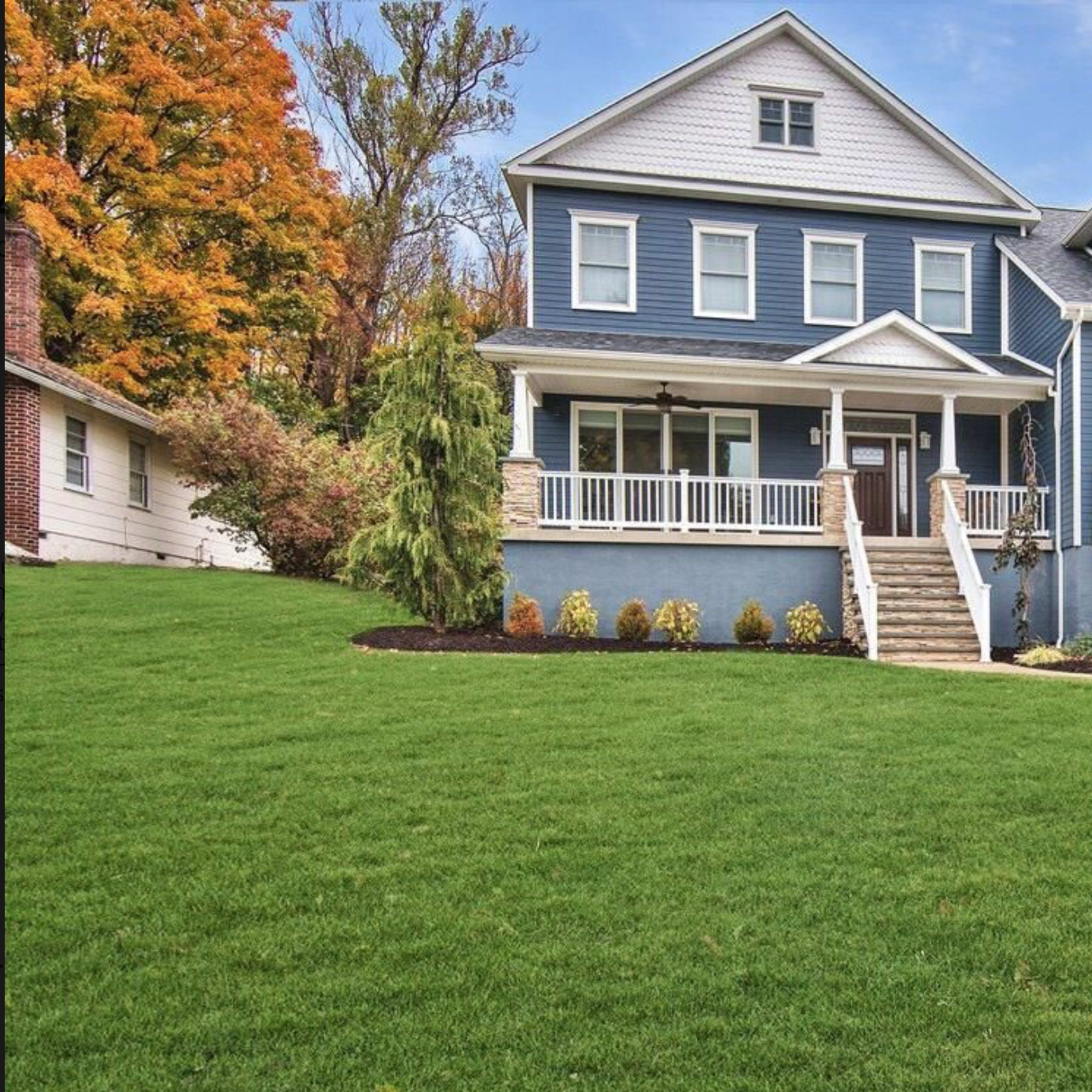 A two-story blue house with white trim and multiple balconies, surrounded by a well-maintained lawn and landscaped shrubs.