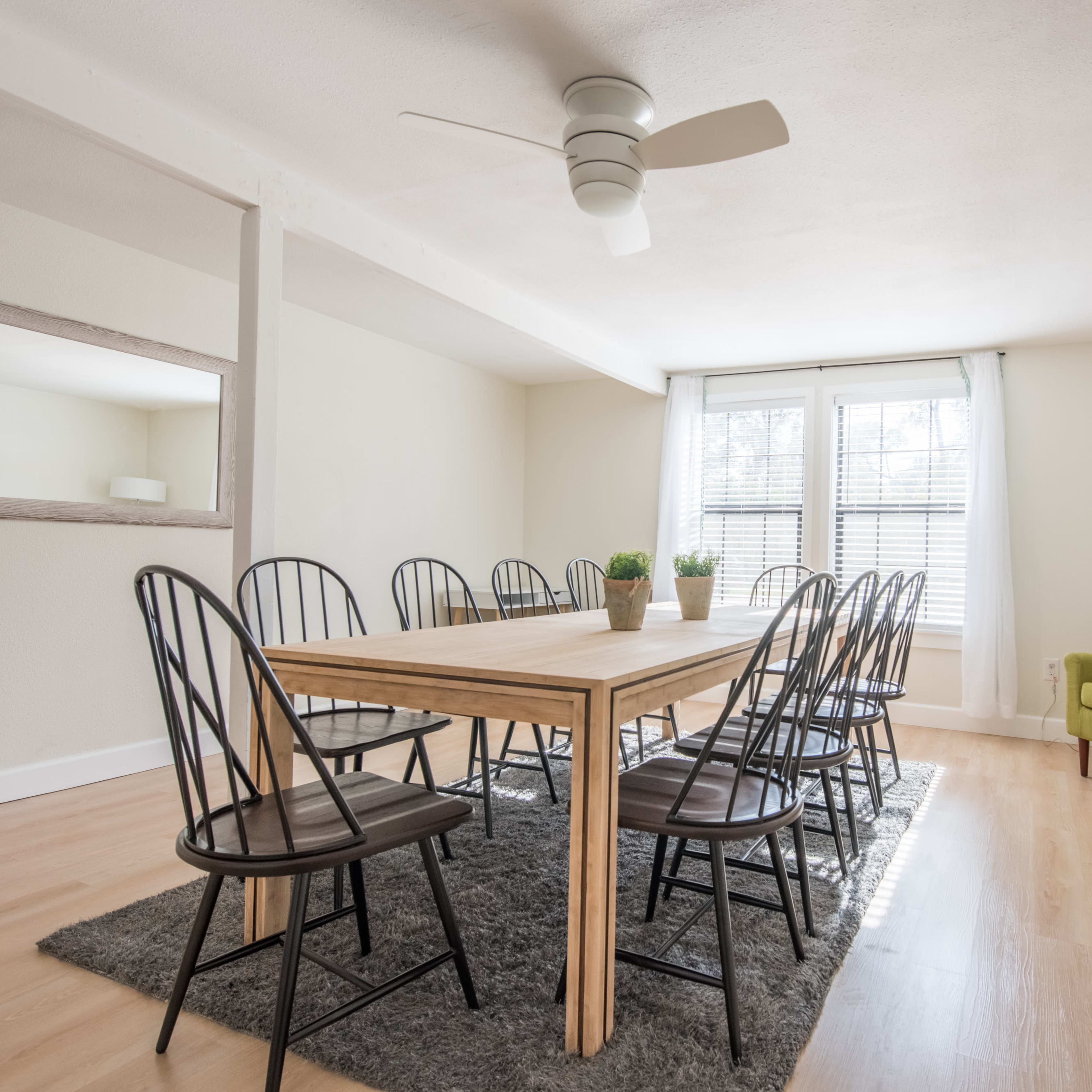 A dining room features a large wooden table surrounded by black chairs, with a green armchair in one corner and a ceiling fan overhead.