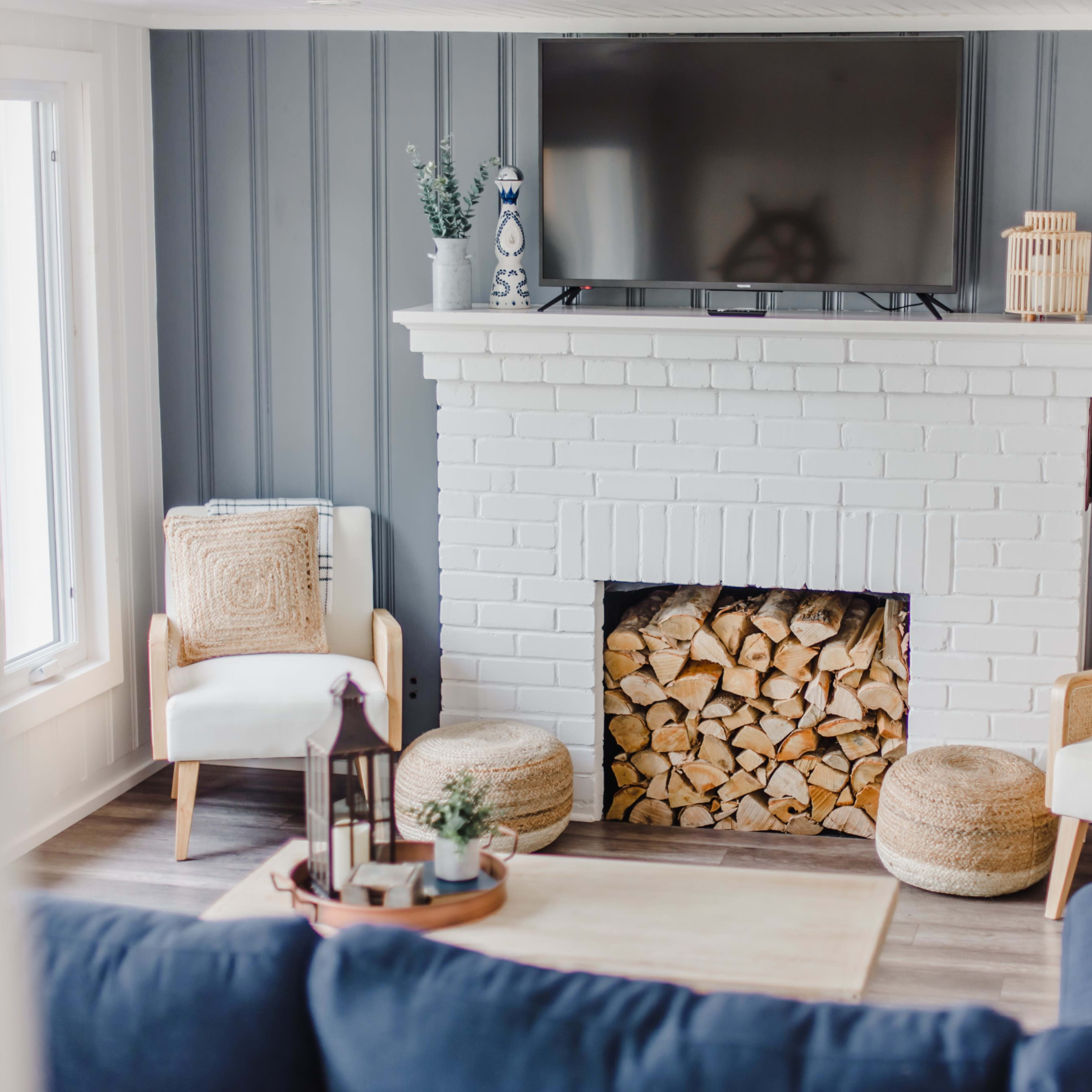 A living room with a white brick fireplace, a flat-screen TV mounted above it, and two light-colored chairs flanking a pile of stacked firewood.