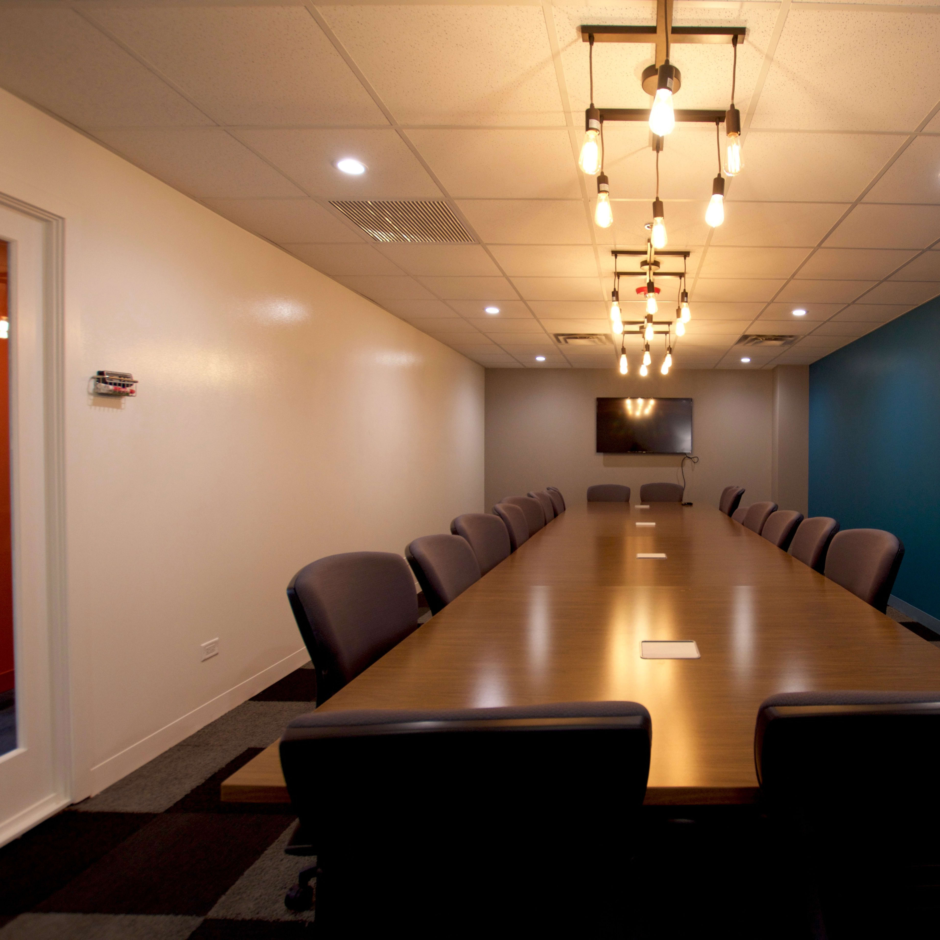 A long conference table with dark chairs is positioned in a well-lit meeting room featuring blue and white walls and a television mounted on one wall.