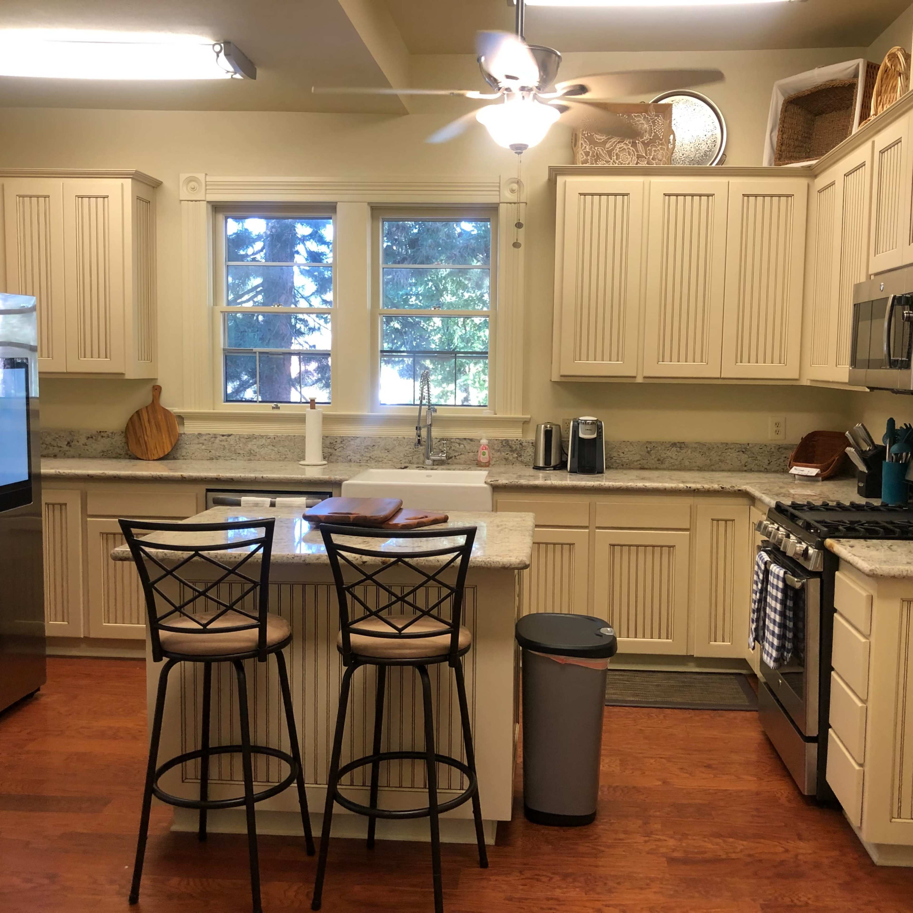 A modern kitchen features light-colored cabinetry, a granite countertop island with two bar stools, and stainless steel appliances.