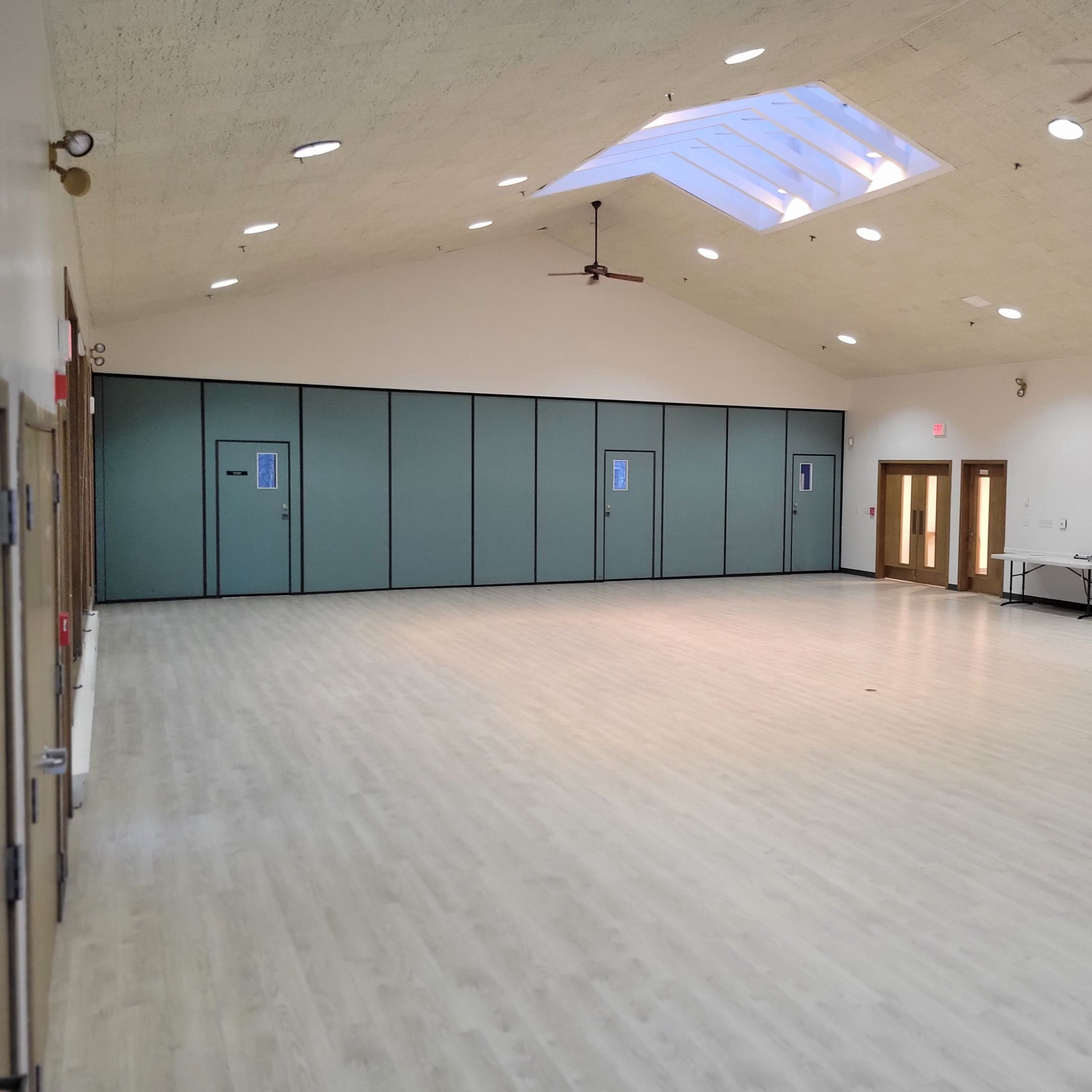 A spacious indoor room with a high ceiling, large windows, and a wood floor, featuring two sets of double doors and a small table against the wall.