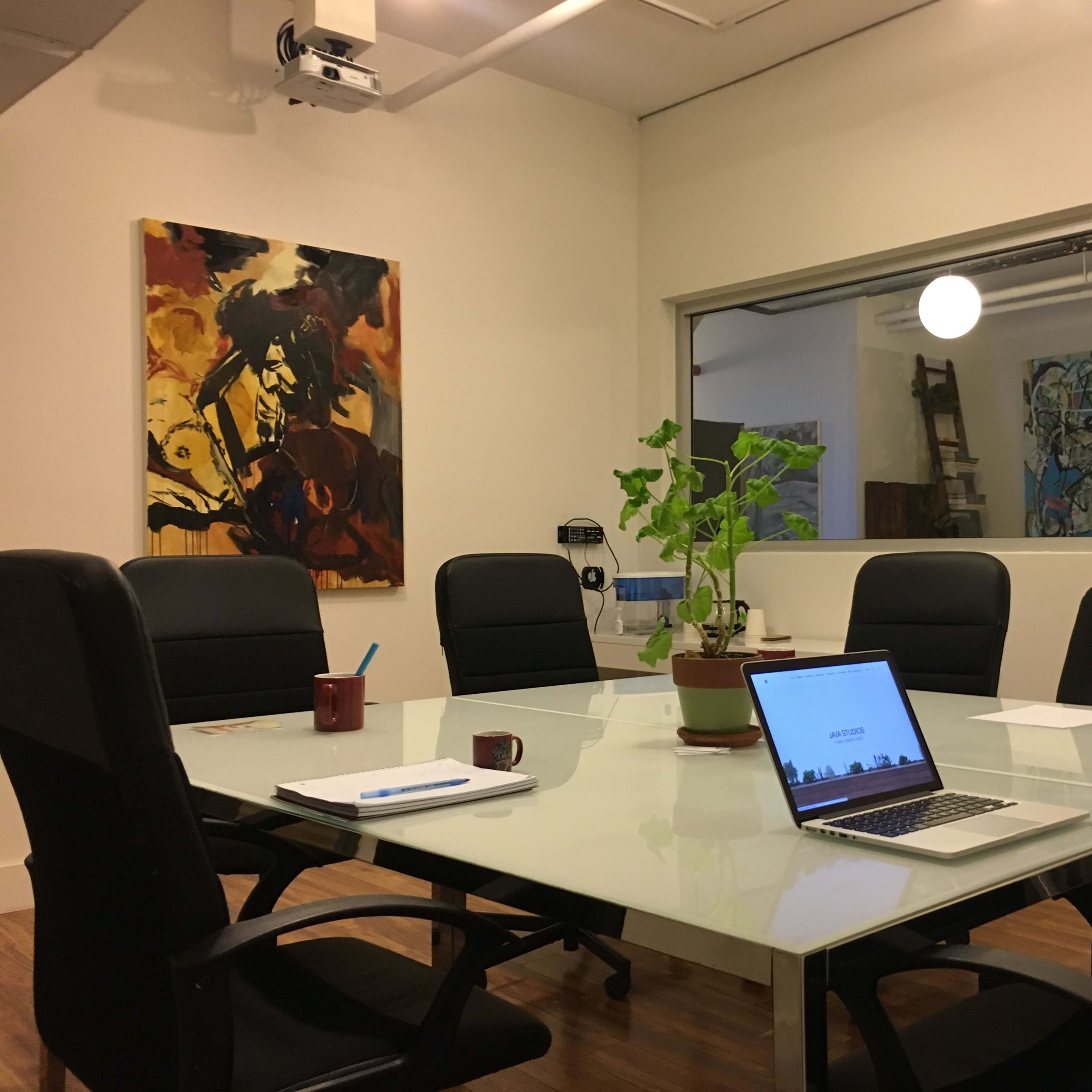 A modern conference room features a glass table surrounded by black chairs, a laptop, a plant in the center, and two artworks on the walls.