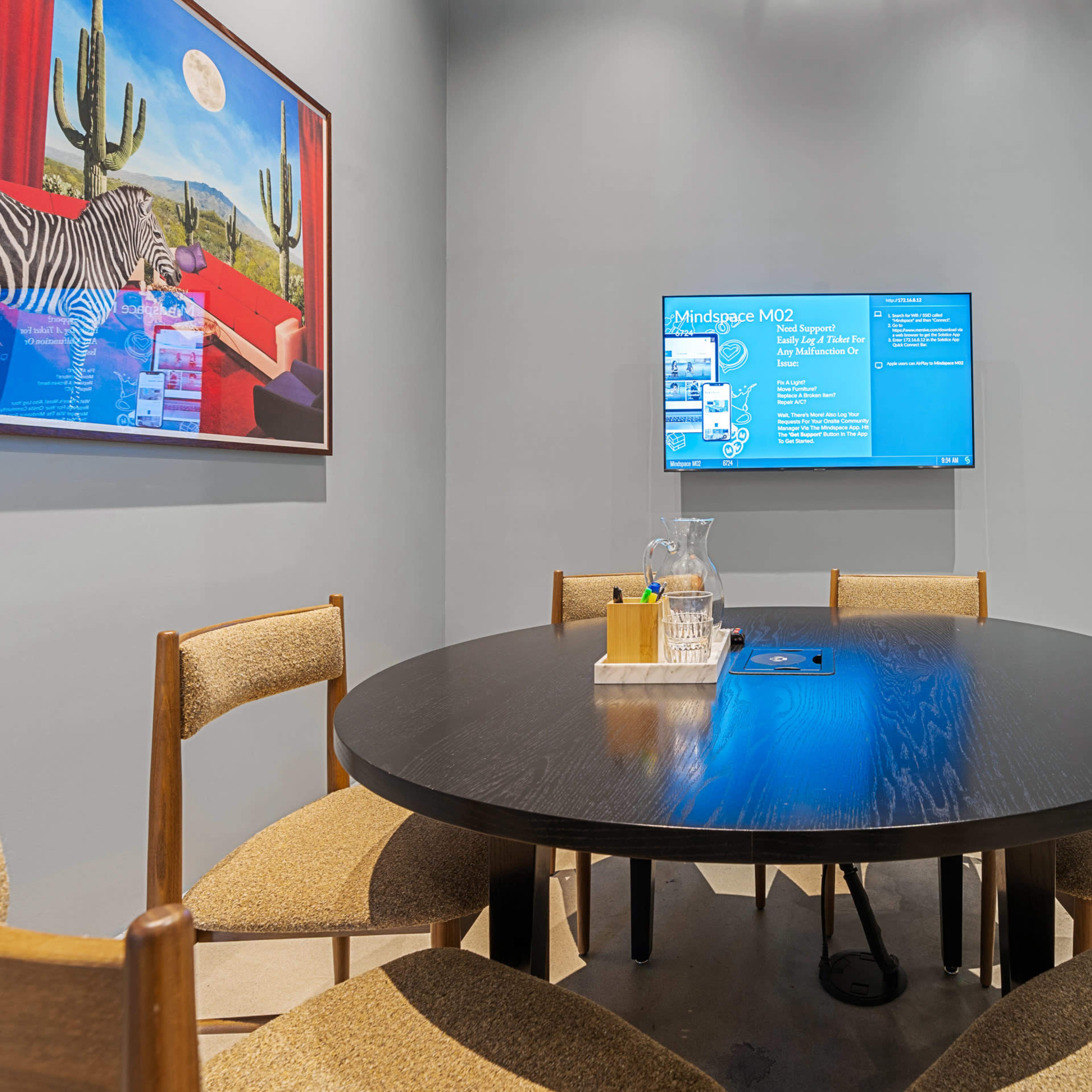 A round table with six chairs is set in a small conference room featuring a wall-mounted screen and artwork of a zebra.