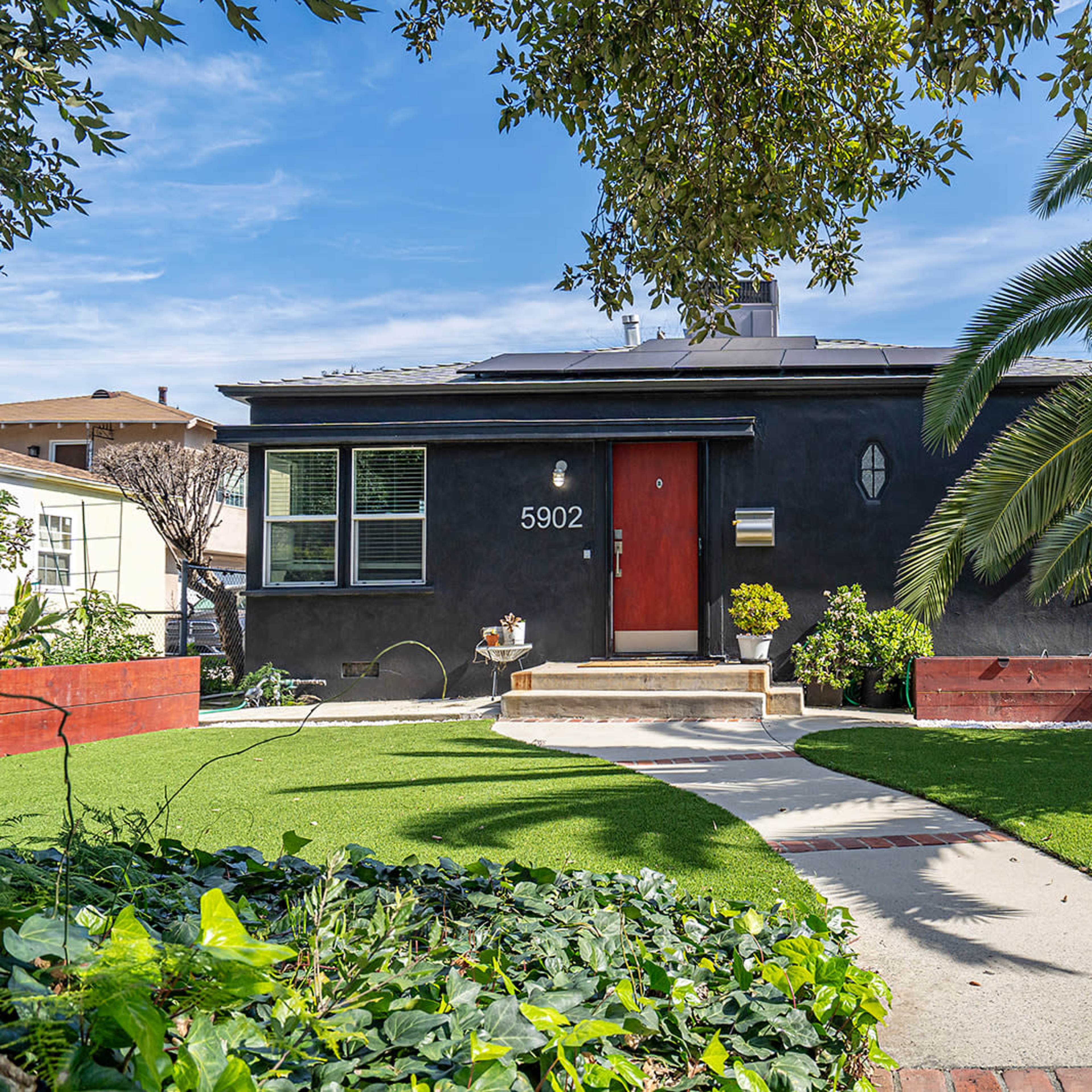 A black house with a red door is surrounded by lush green grass, palm trees, and garden beds.