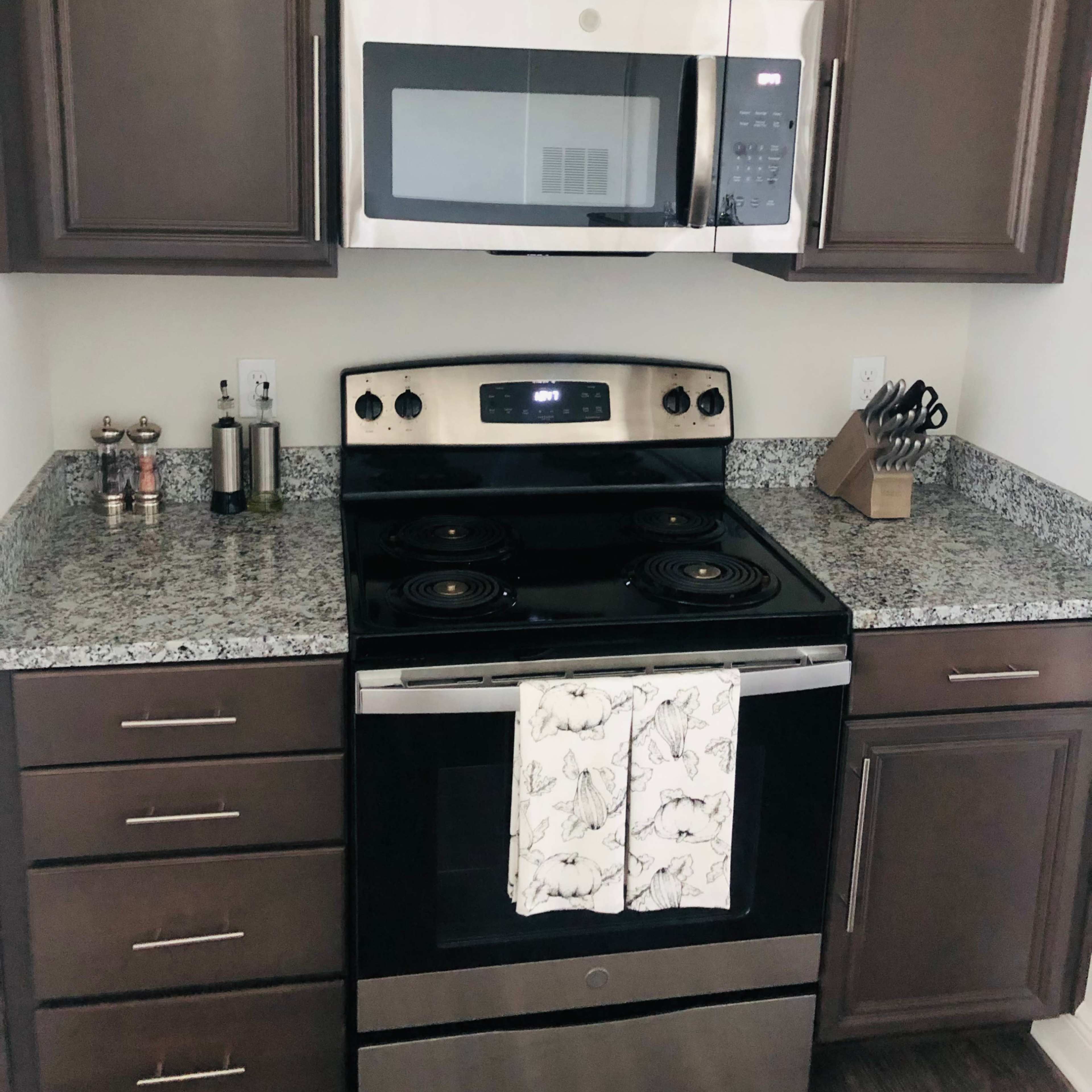 A modern kitchen corner with a stainless steel stove and microwave above it, surrounded by dark cabinets and a speckled granite countertop.