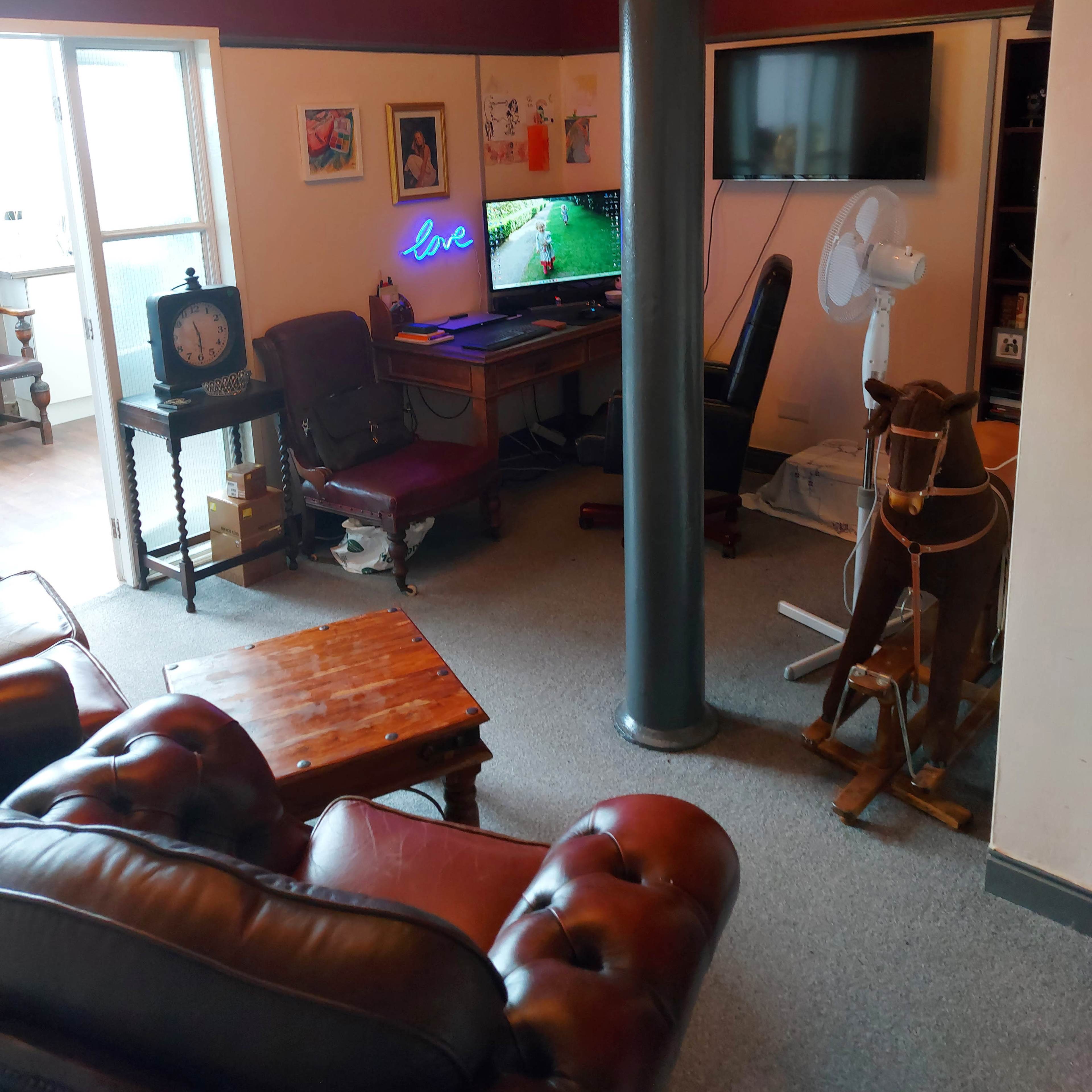 A cozy room with leather seating, a desk with a television, and various artworks on the walls, including a portrait and a neon sign that reads "love."