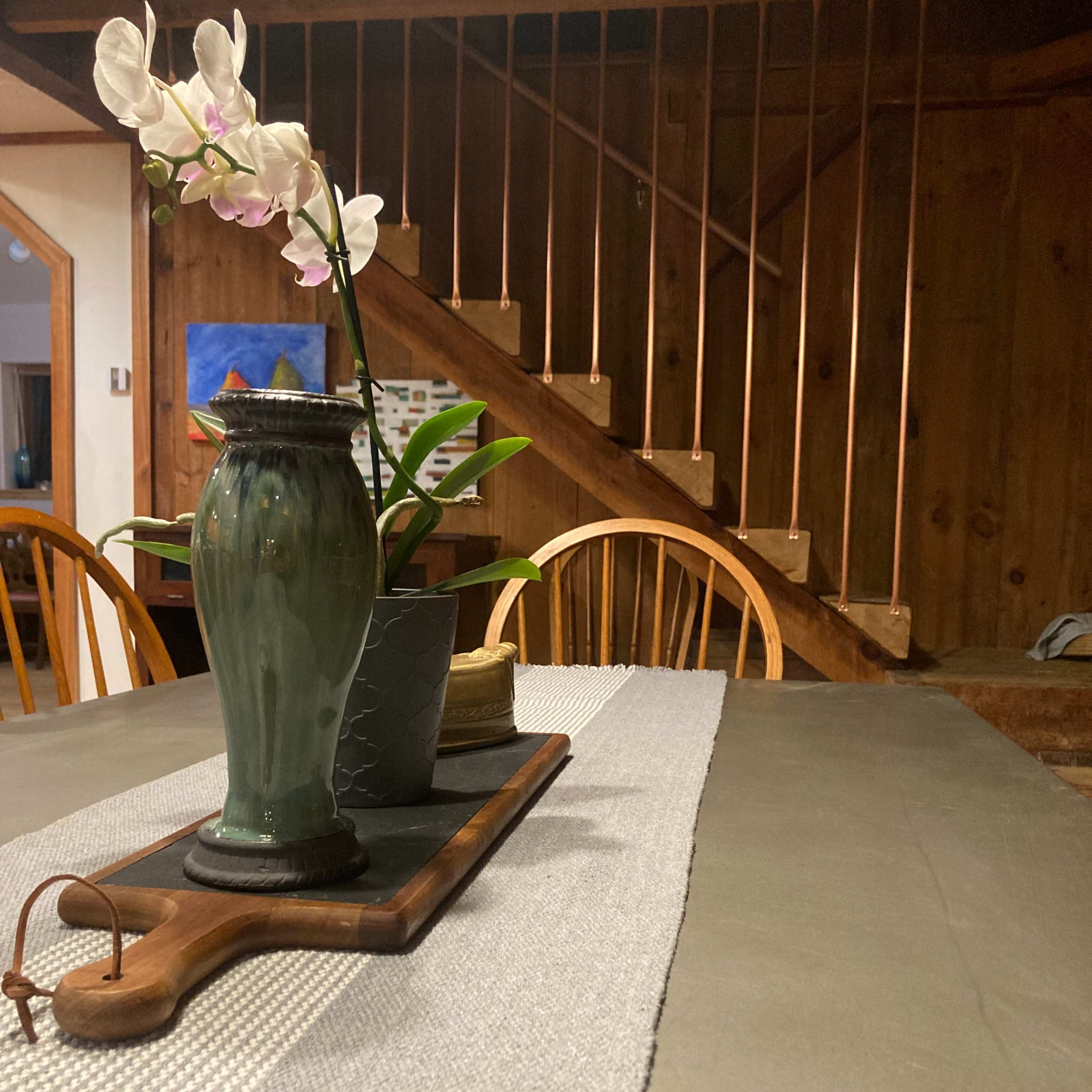 A dining table is set with a decorative vase containing an orchid, a wooden cutting board, and a textured runner in a rustic interior with a staircase in the background.
