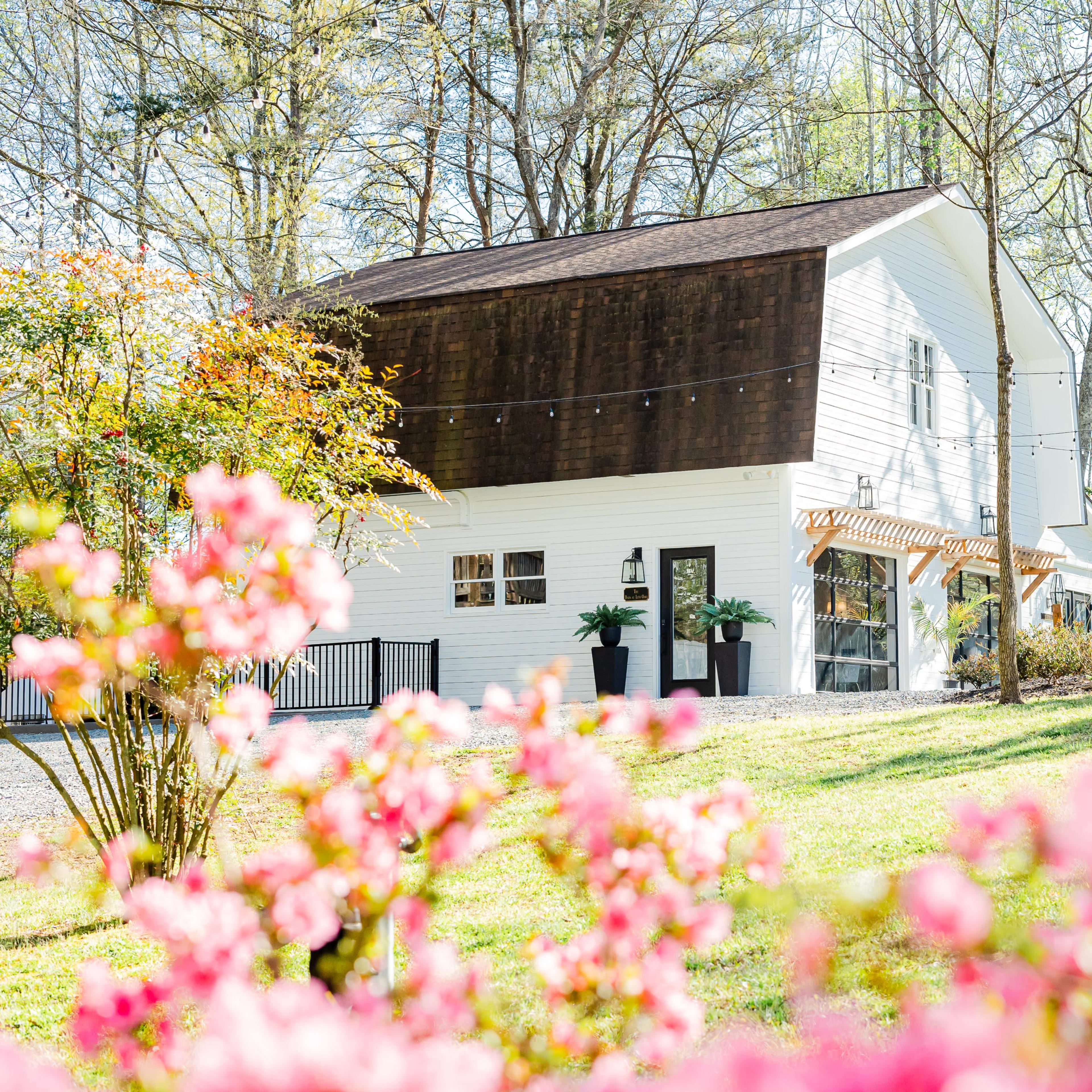 A white building with a dark wooden upper section is nestled among trees and surrounded by blooming pink flowers.