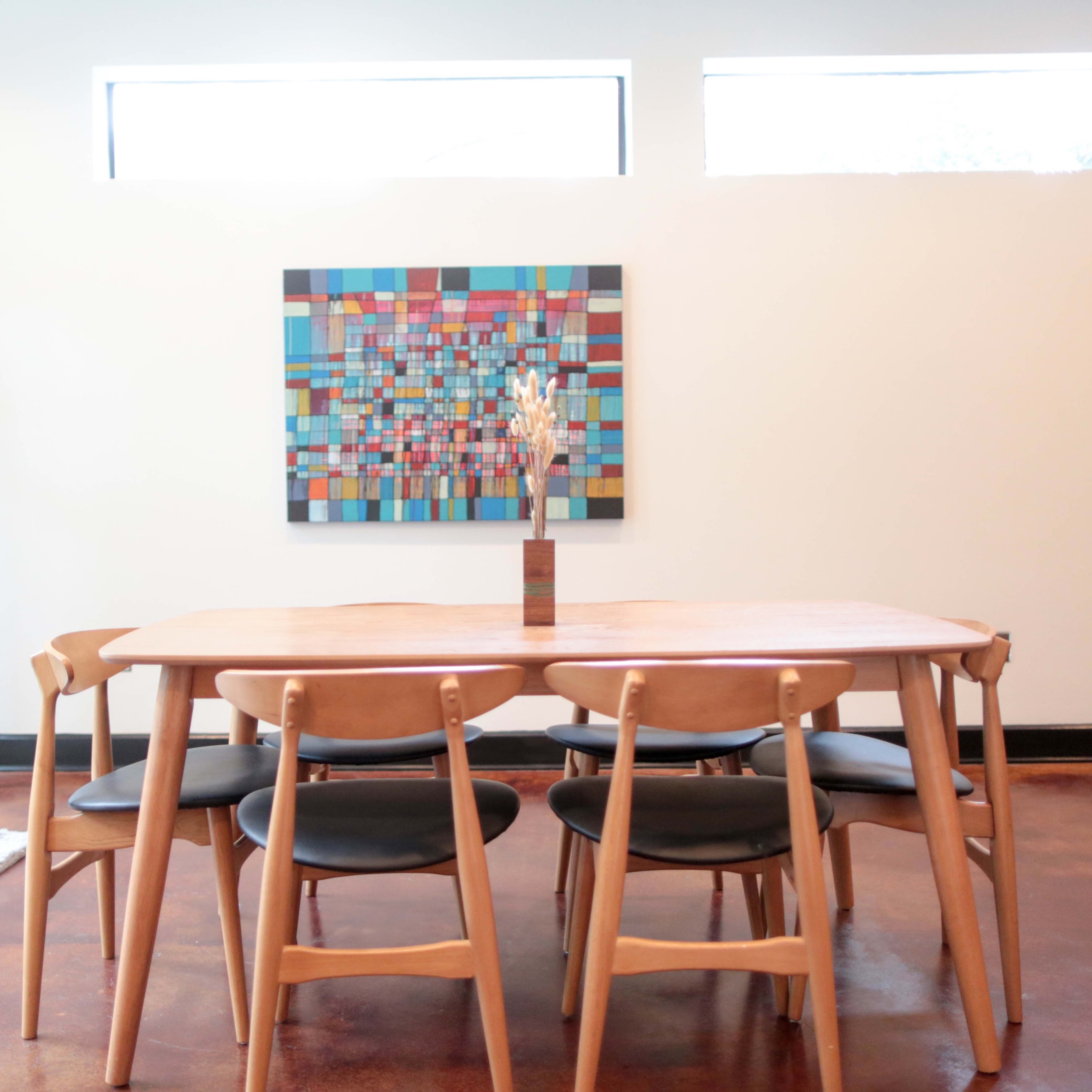 A wooden dining table with black chairs is set in a room featuring a colorful abstract painting and a display case.