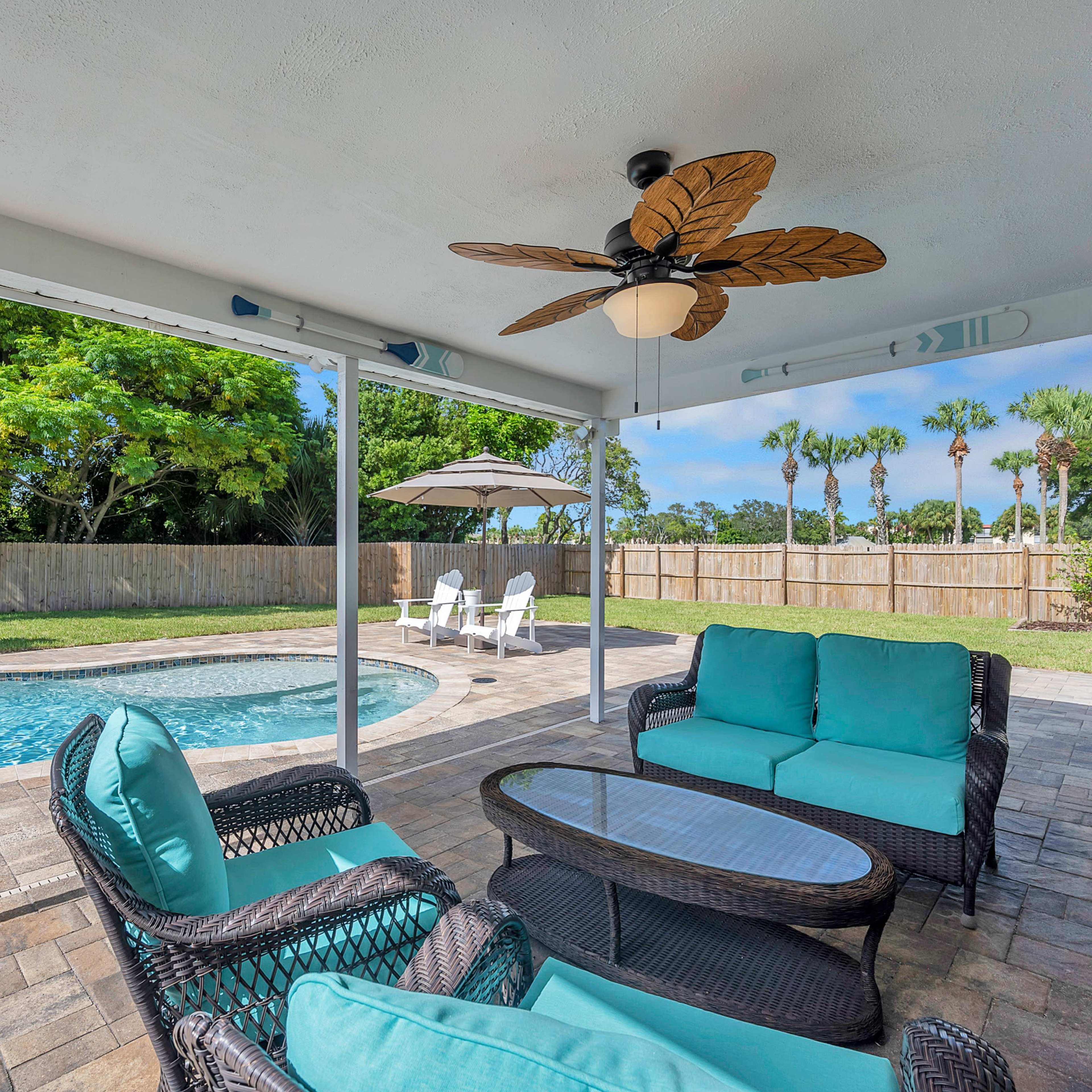 A covered patio area with wicker furniture and a ceiling fan overlooks a pool surrounded by a paver deck and tropical landscaping.