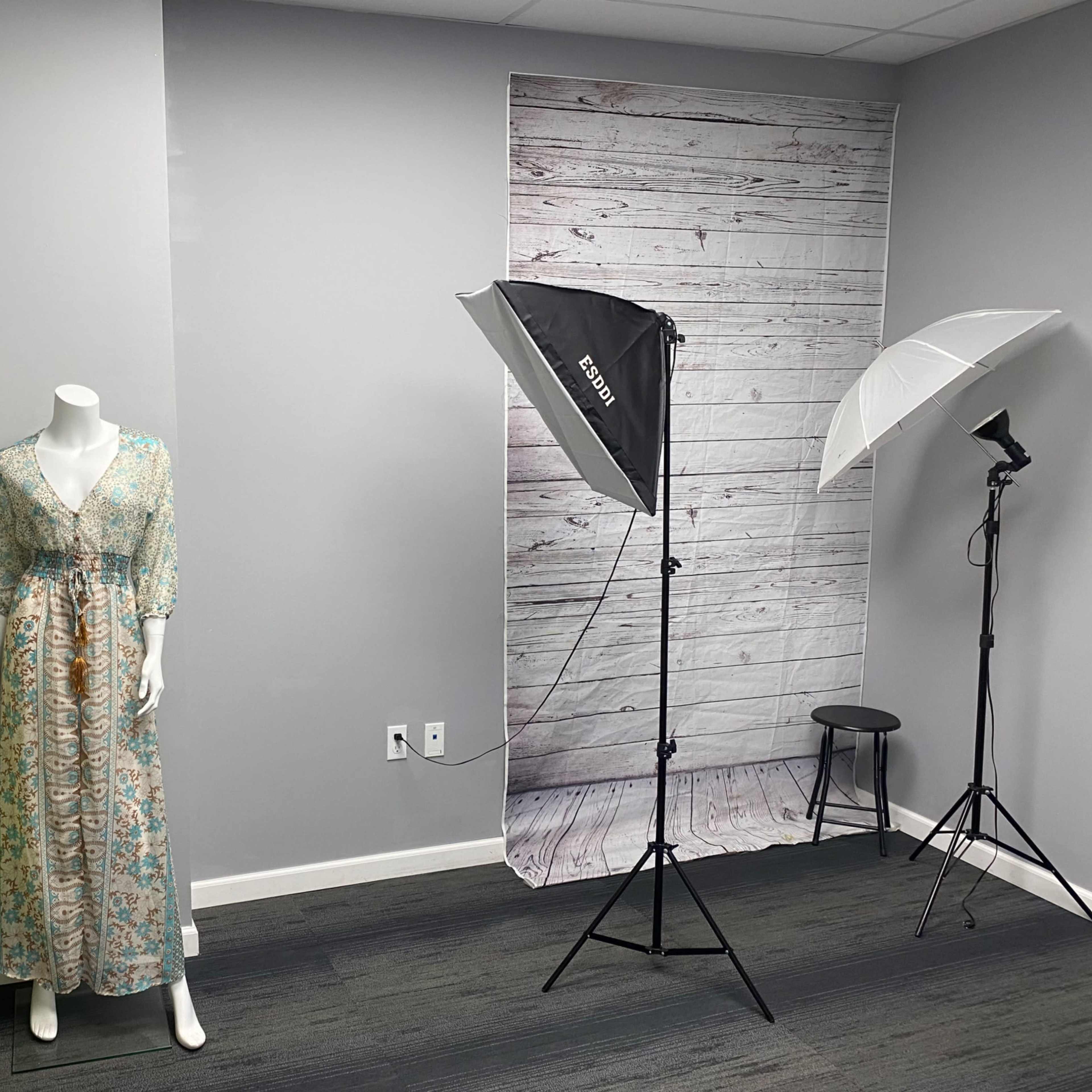 A mannequin wearing a floral dress stands next to photography lighting equipment in a room with gray walls and a wood-patterned backdrop.