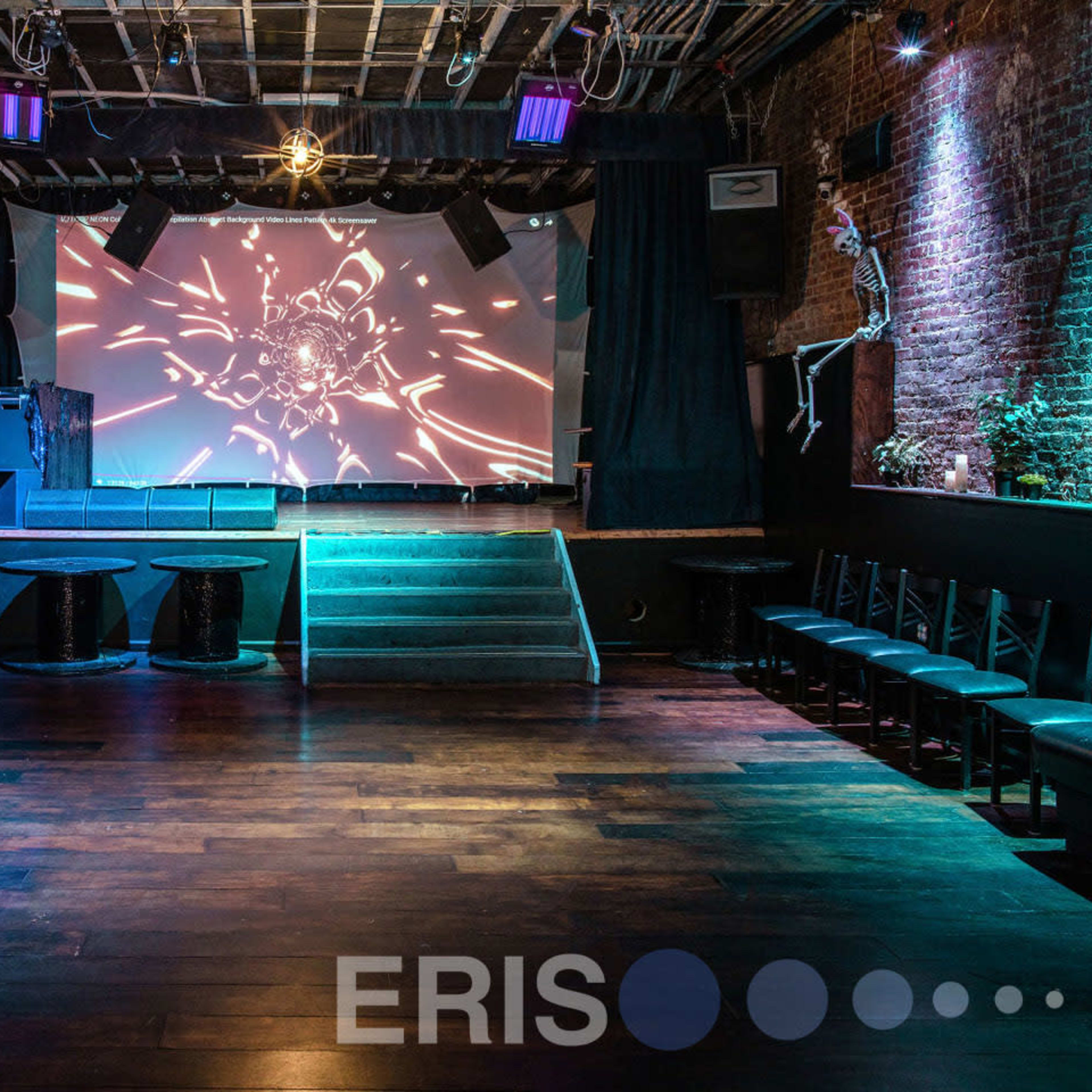 An empty music venue with a stage displaying visual projections, surrounded by seating areas and dim lighting.