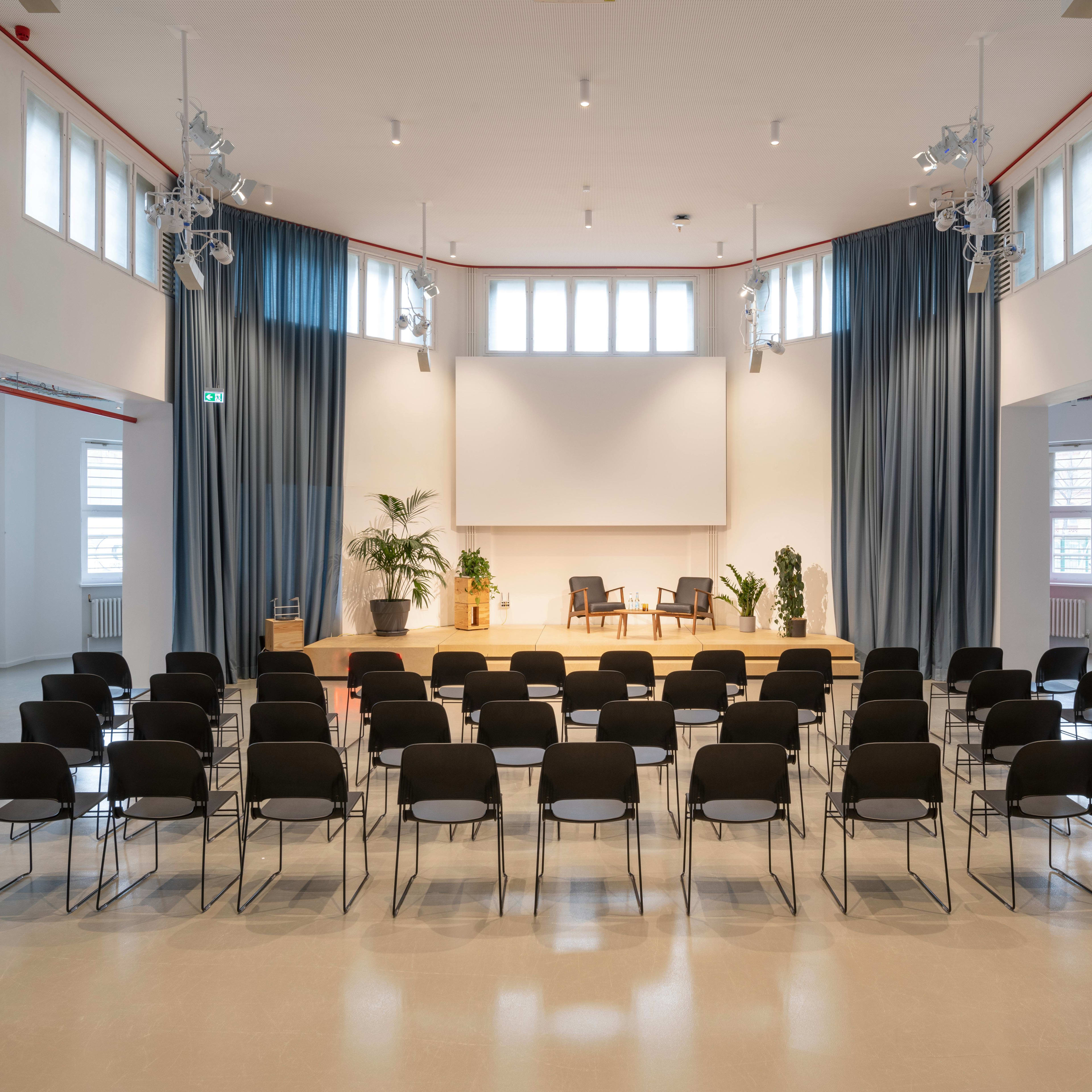 A spacious indoor venue set up with rows of black chairs facing a wooden stage that features two chairs and a large screen, with curtains and plants decorating the surrounding area.