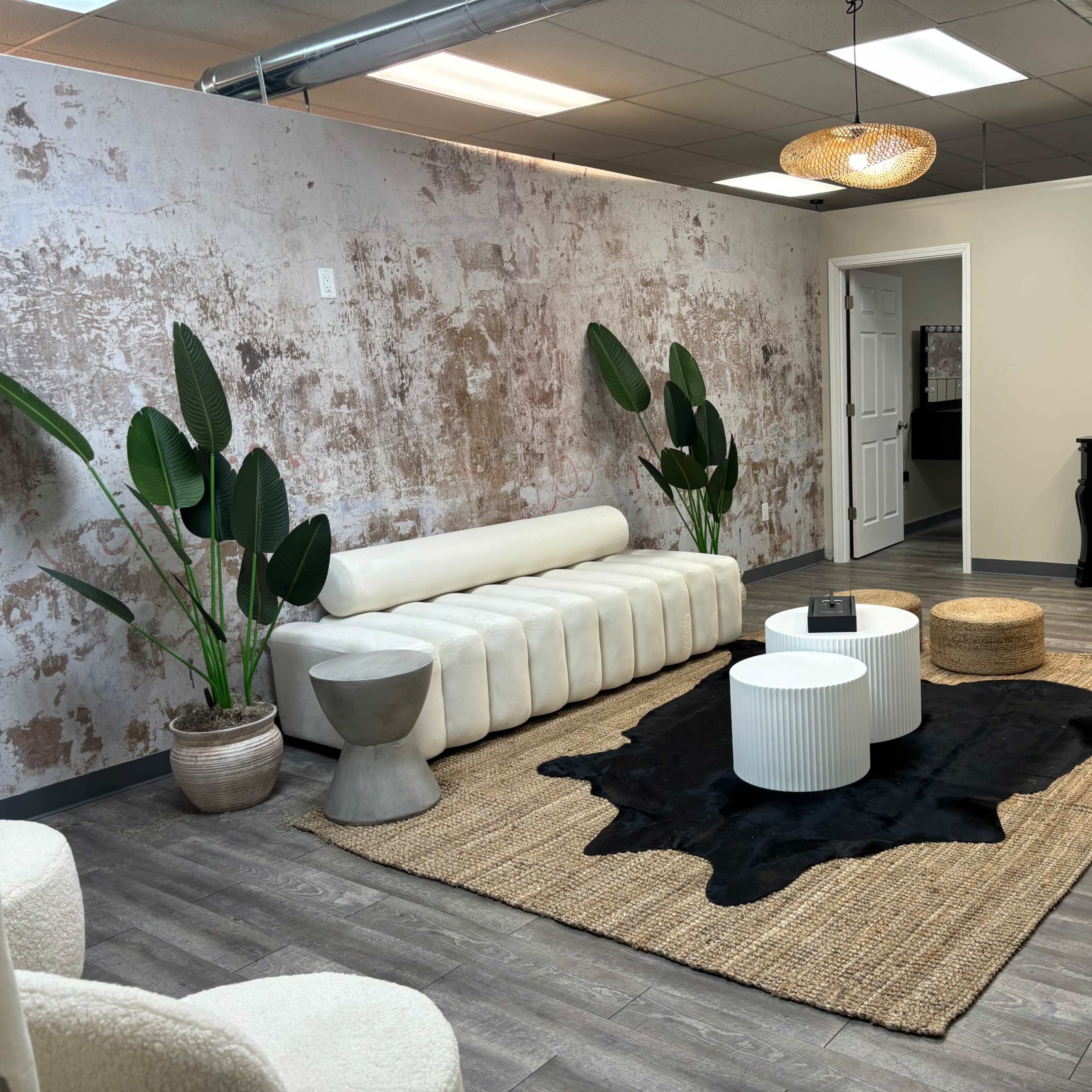 A modern interior space with a light-colored sofa, several round coffee tables, a woven rug, and decorative plants against a textured wall.