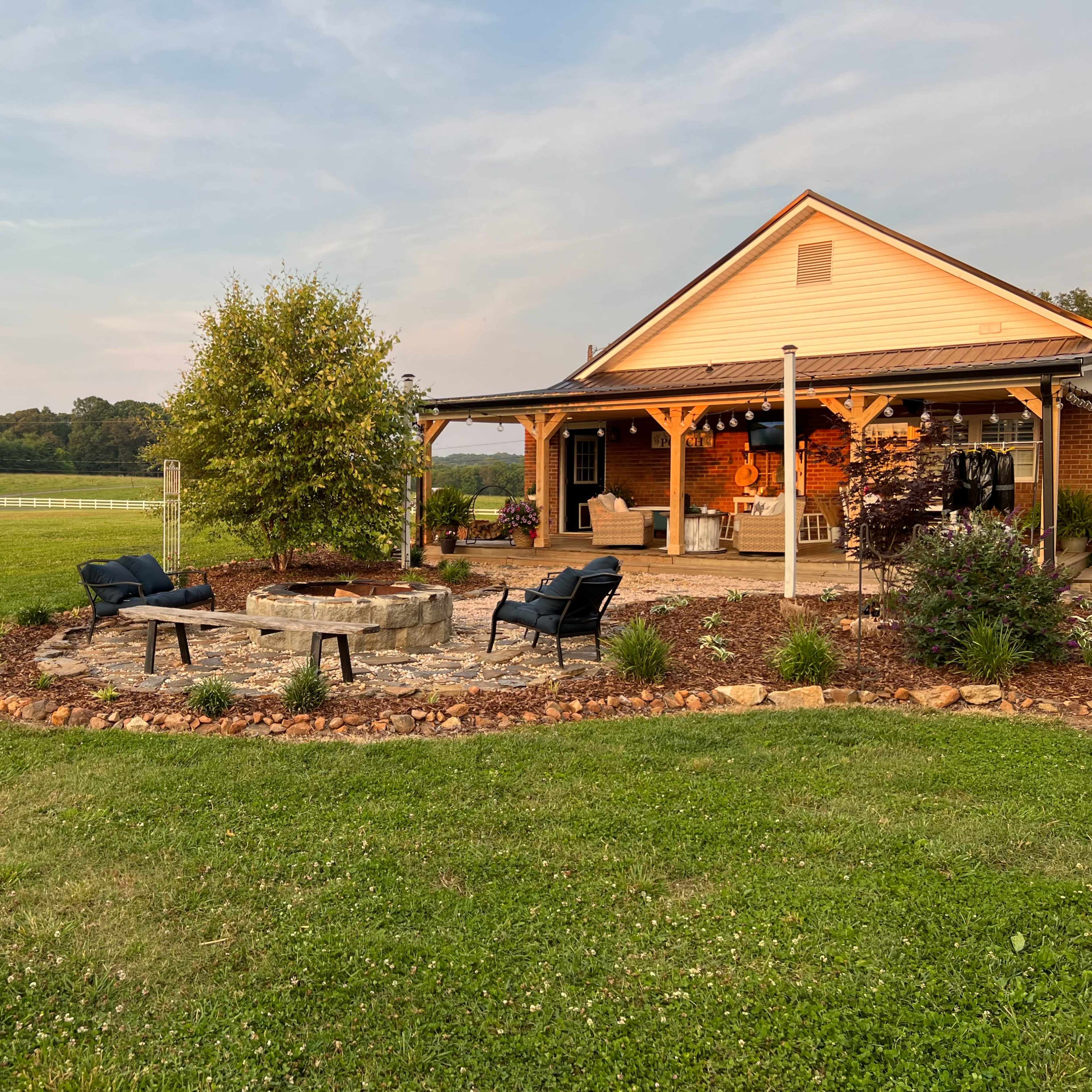 A cozy house with a stone fire pit and seating area is surrounded by a lush green lawn and trees.