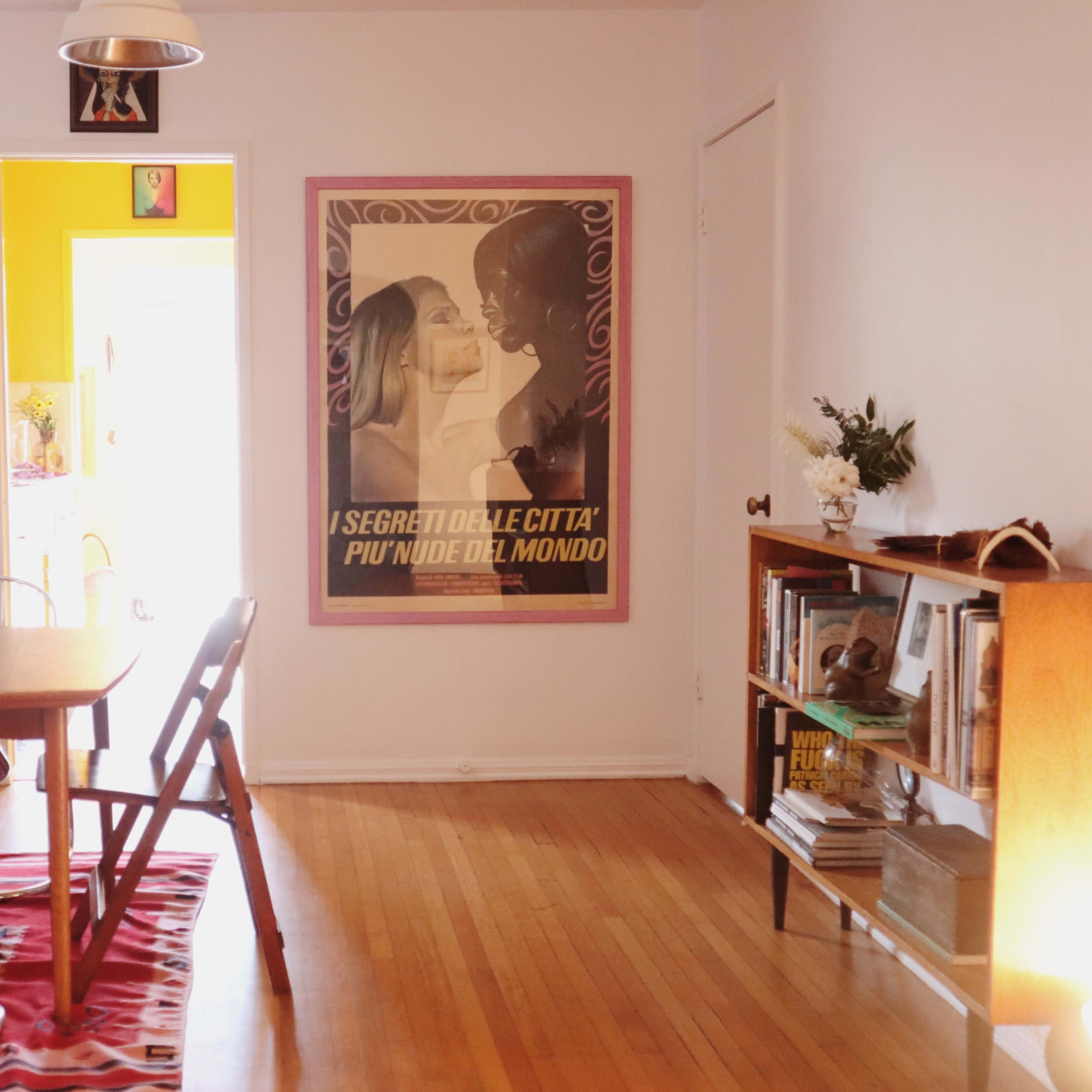 A cozy room featuring a wooden dining table surrounded by chairs, complemented by a large poster on the wall and a bookshelf nearby.