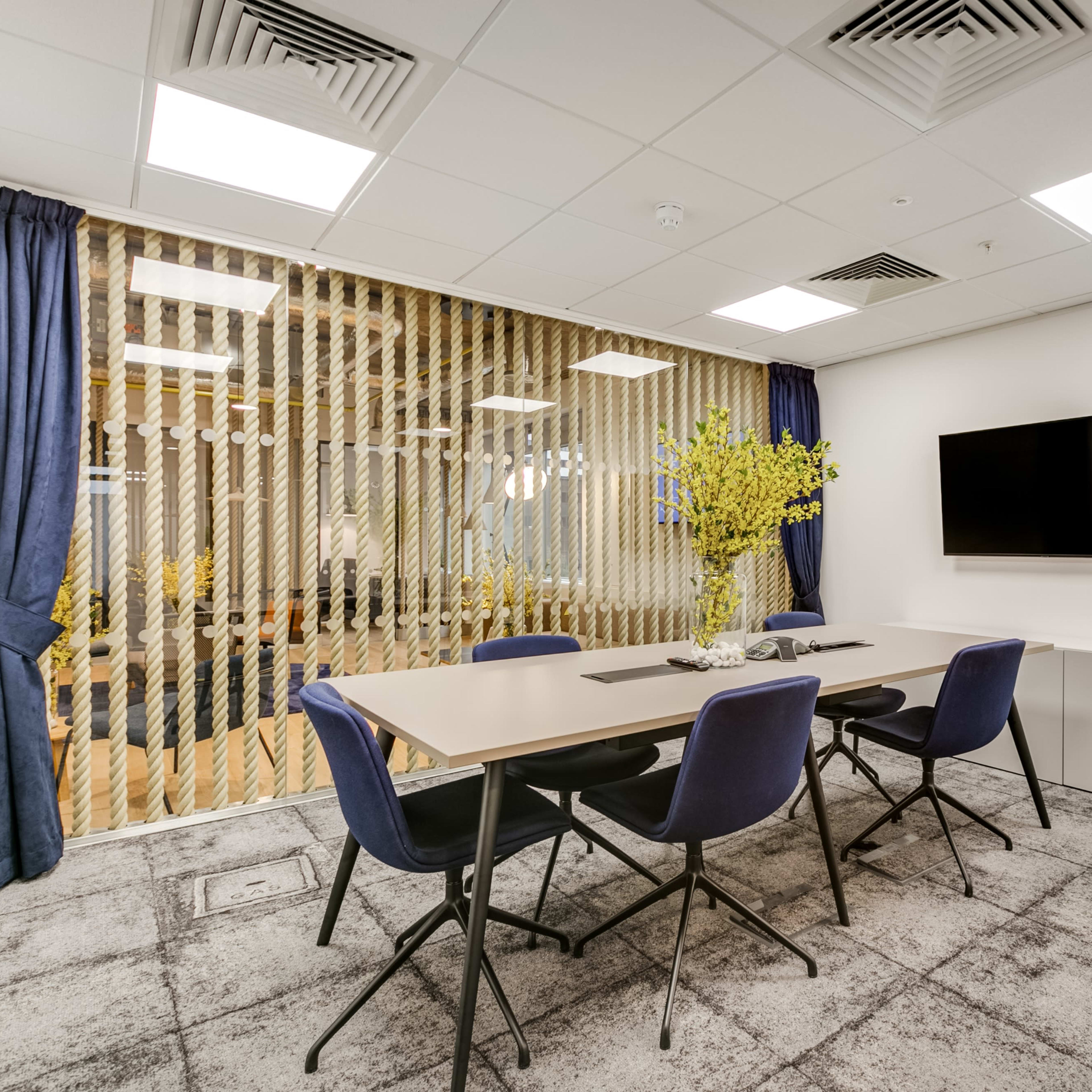 A meeting room features a large table with four chairs, a wall-mounted TV, and a window covered with vertical blinds that offers a view of a light-filled space beyond.