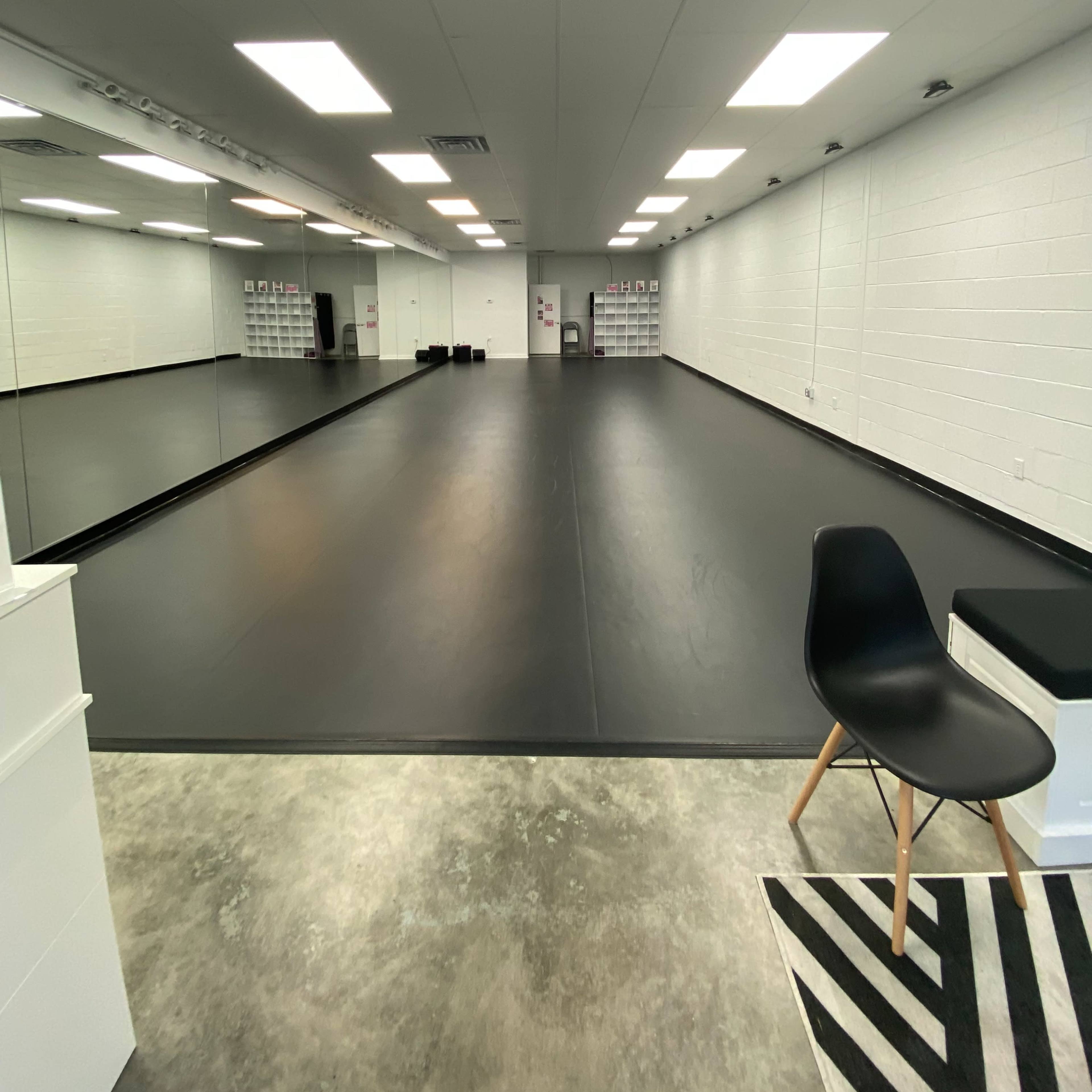 A large dance studio with a black floor, white walls, and bright overhead lighting.