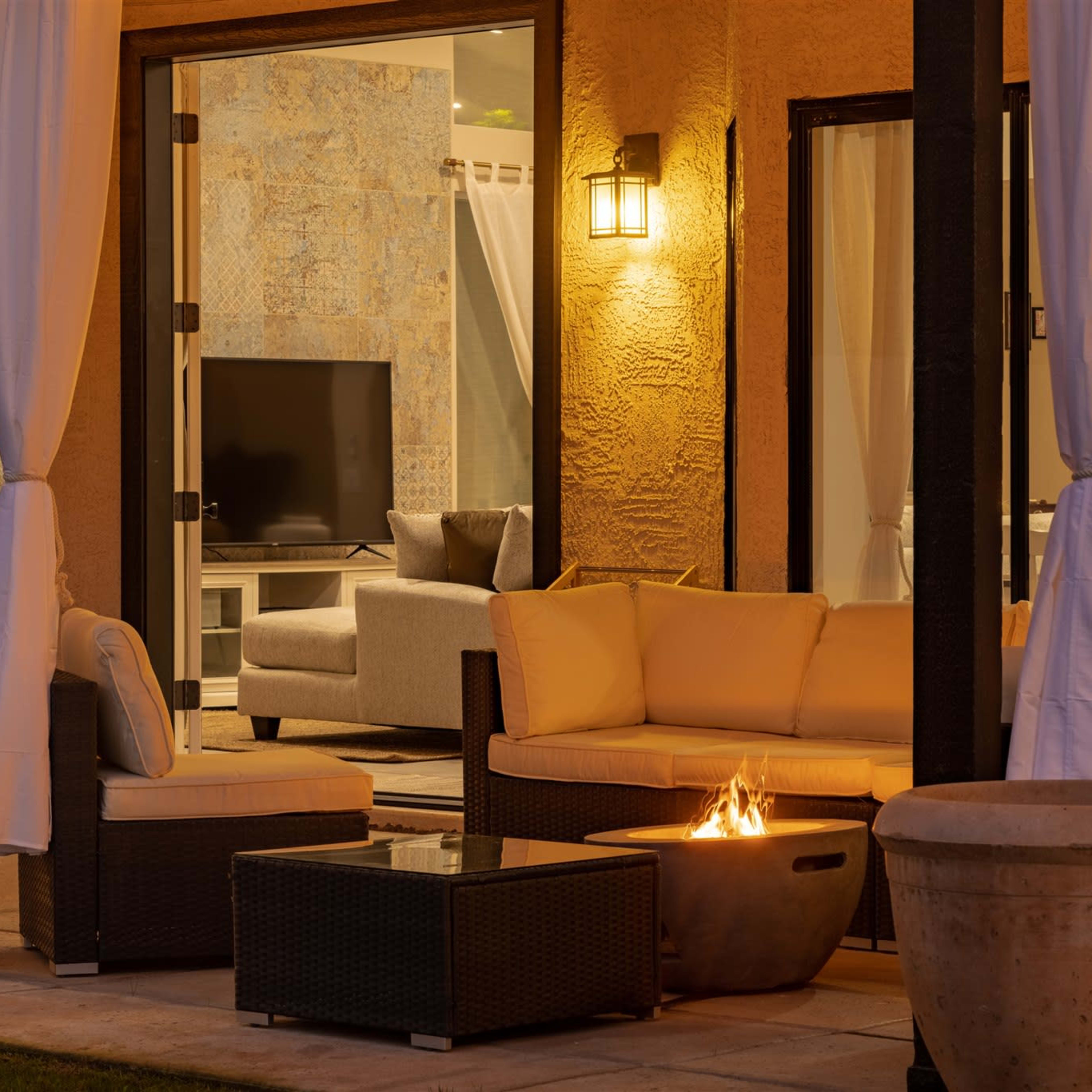 A cozy outdoor seating area features a modern sofa set, a fire pit, and a view of a living room through an open sliding door.