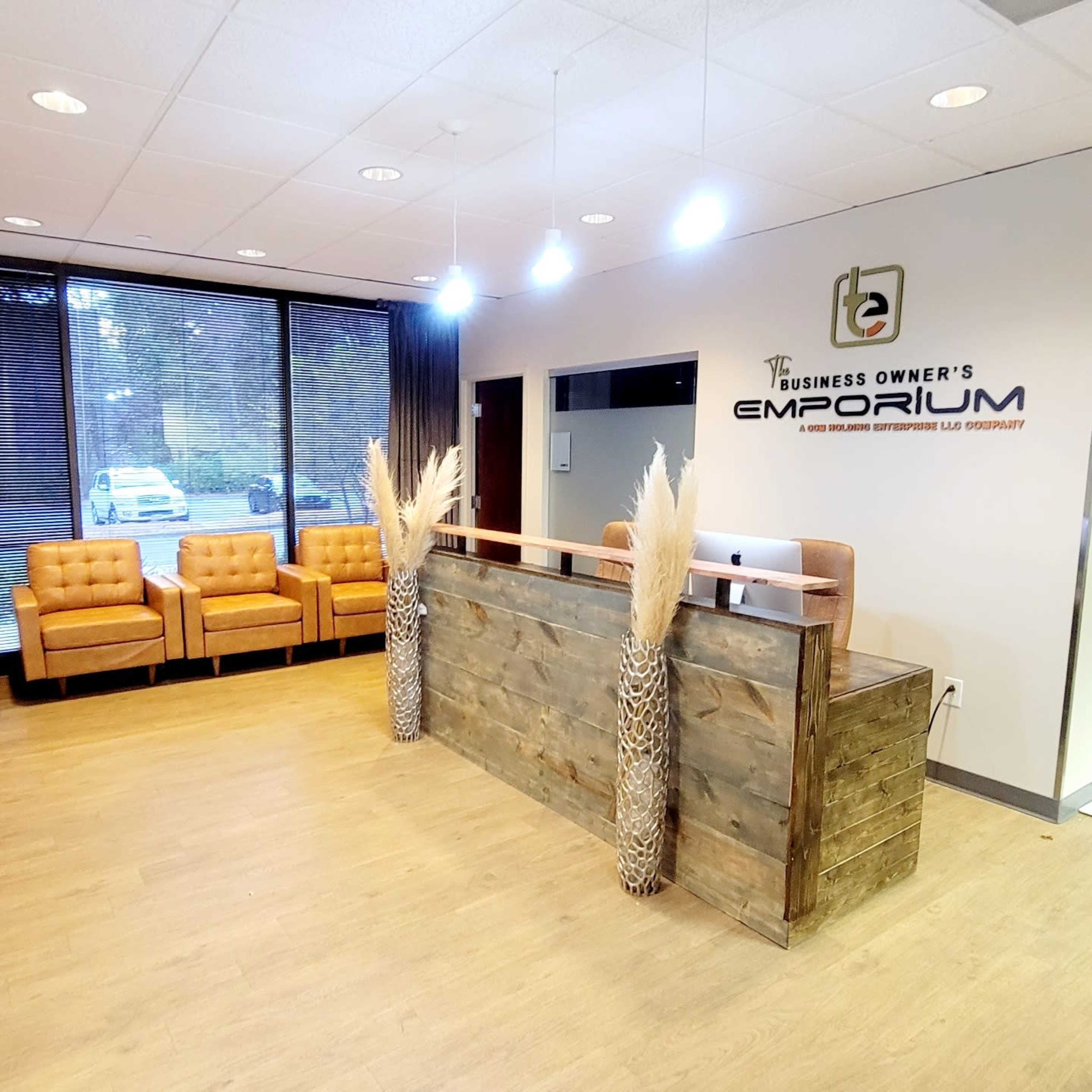A reception area featuring brown leather chairs, a wooden desk with the sign "Business Owner's Emporium," and large windows with natural light.