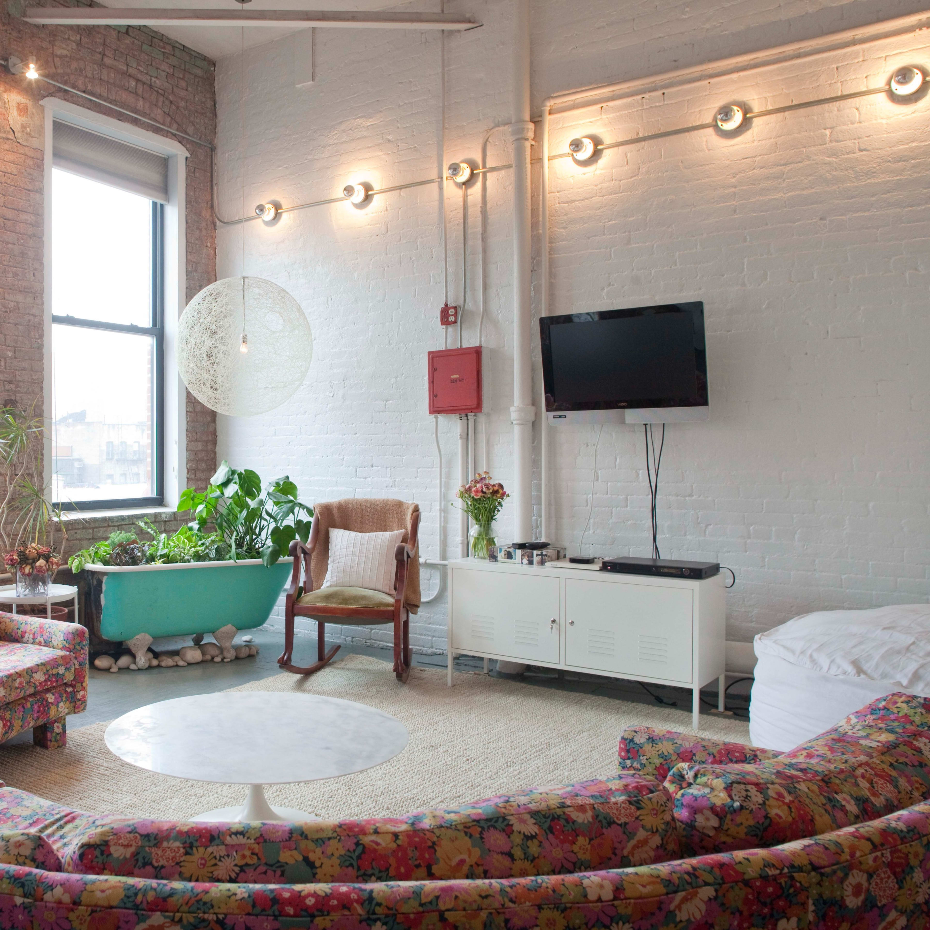A living room features a curved floral-patterned sofa, a round table, a hanging light fixture, and large windows with plants in a blue tub.