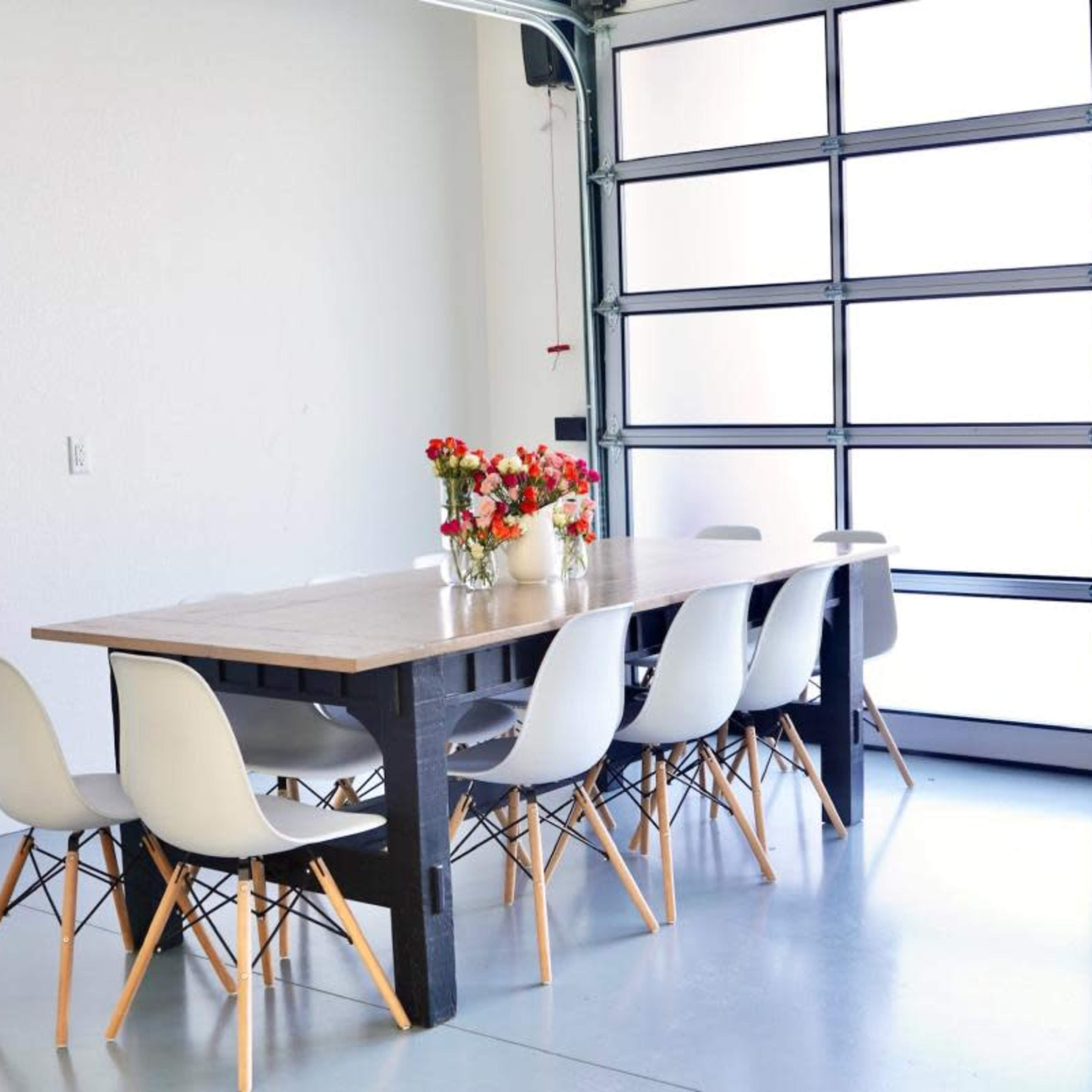 A long wooden dining table with a vase of flowers is surrounded by eight white chairs in a garage with large glass doors.