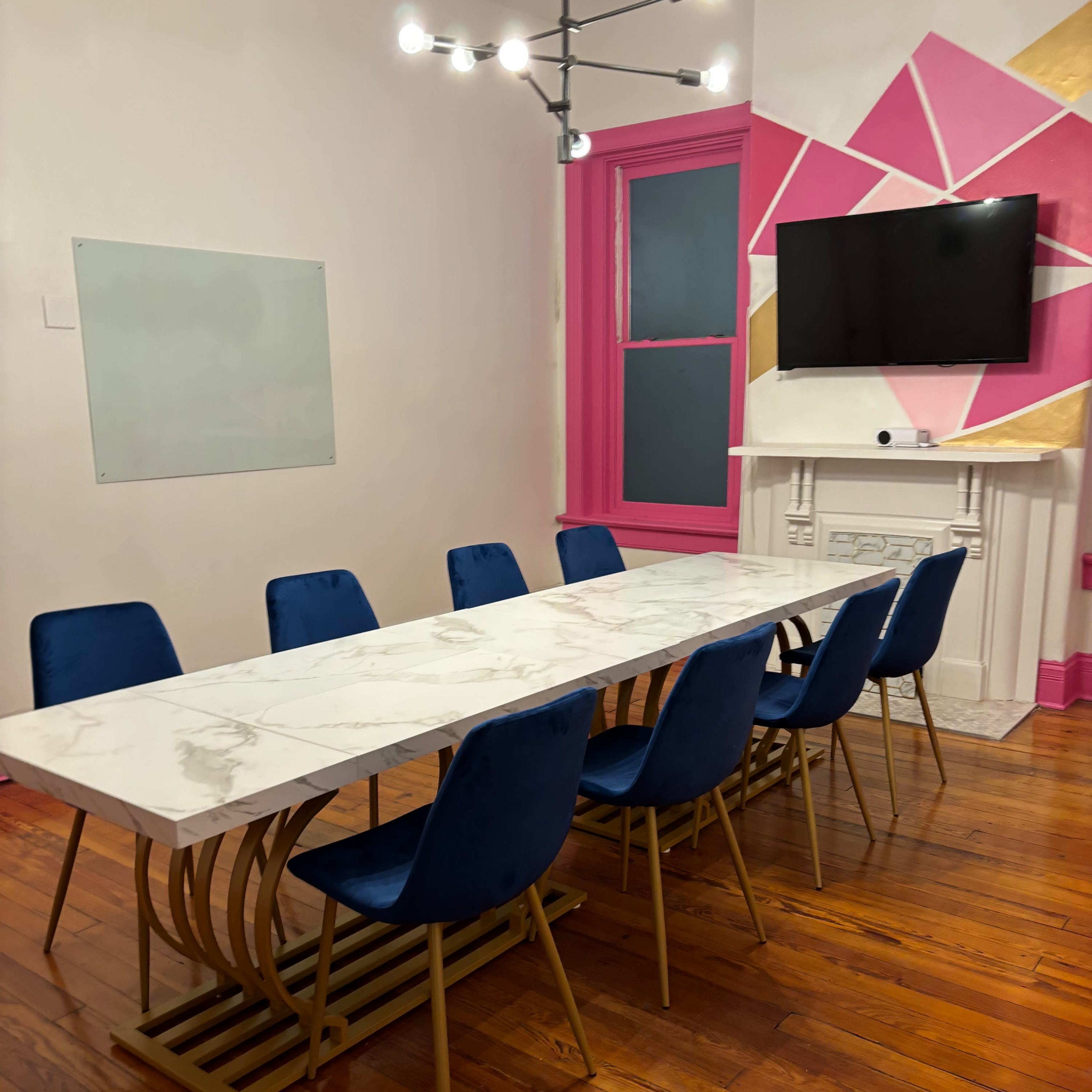 A long marble dining table is surrounded by blue chairs in a brightly colored room with a television mounted on the wall.