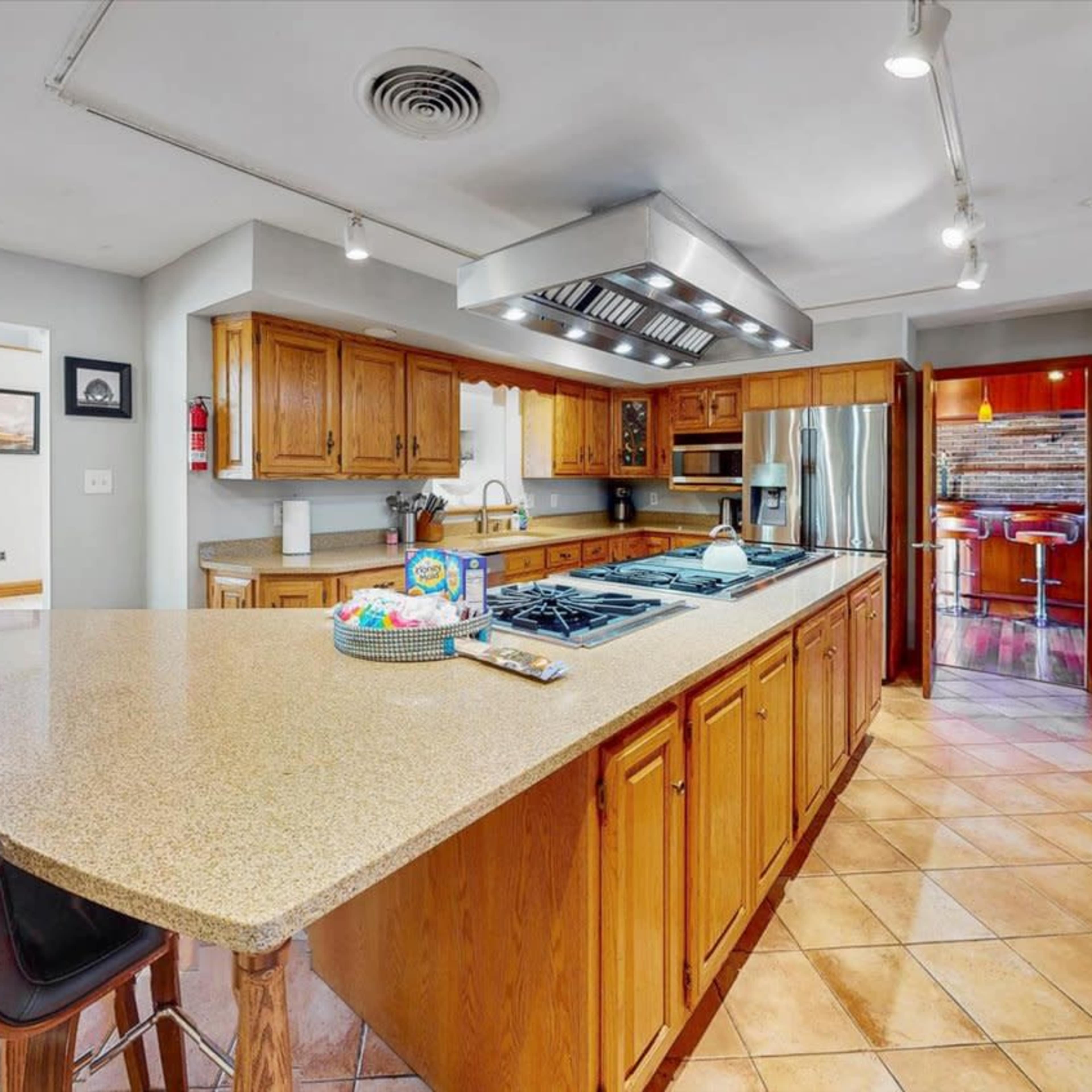 A spacious kitchen with wooden cabinets, a large island with bar seating, a stovetop, and an open view to a dining and pool area.