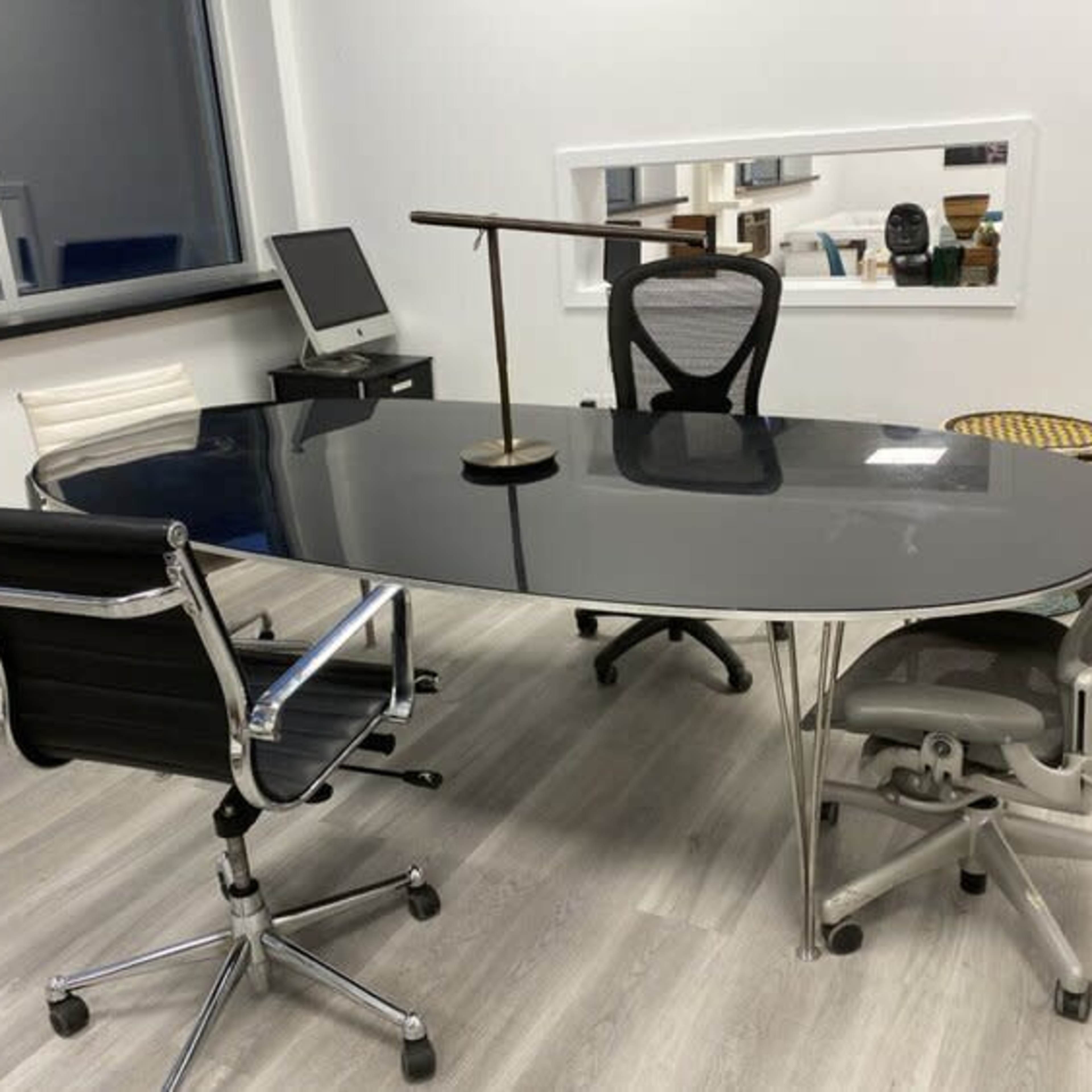 A modern office meeting room features an oval black table surrounded by three ergonomic chairs, with a lamp and a computer setup visible in the background.
