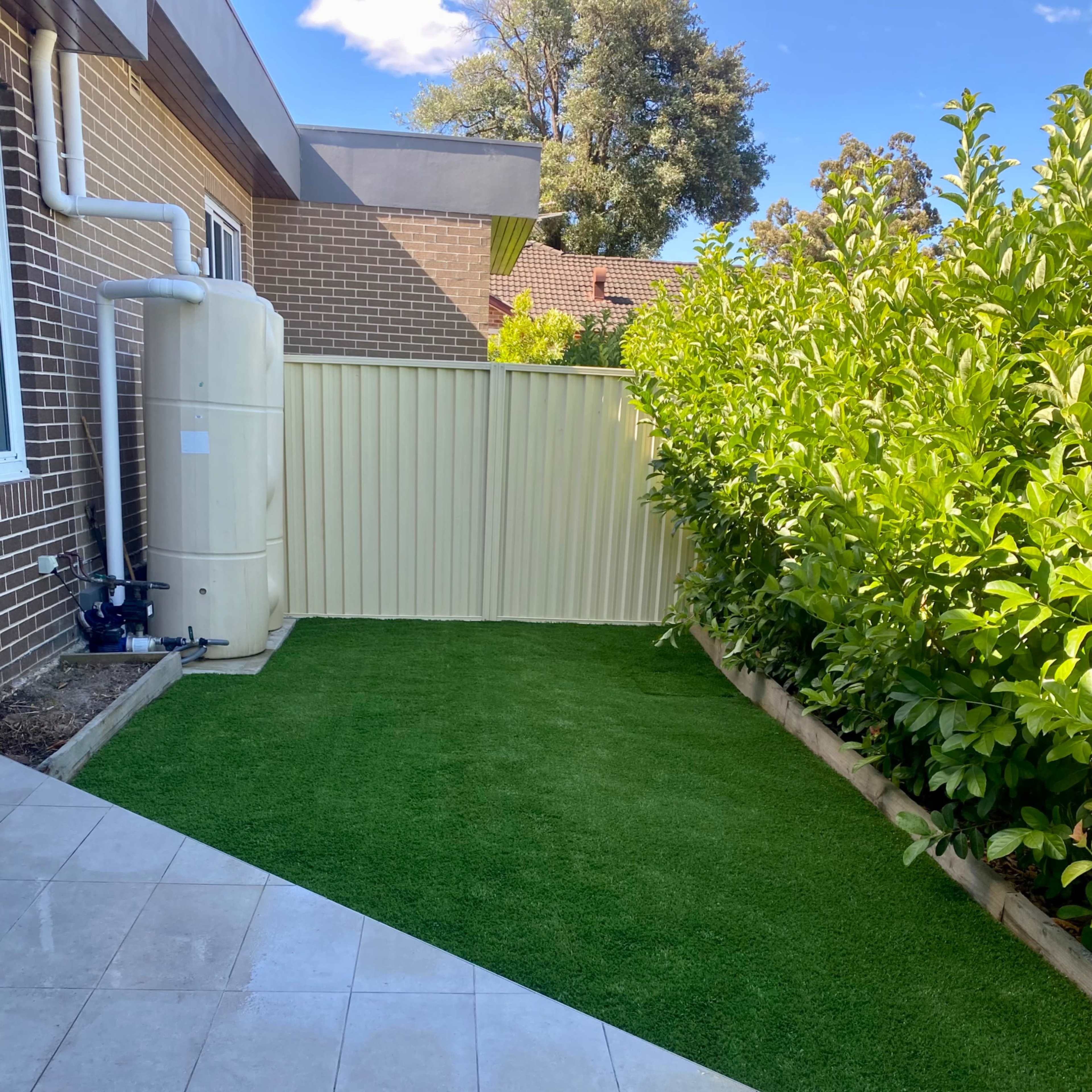 A backyard featuring a section of artificial grass bordered by a fence and a house, with a water tank visible near the wall.
