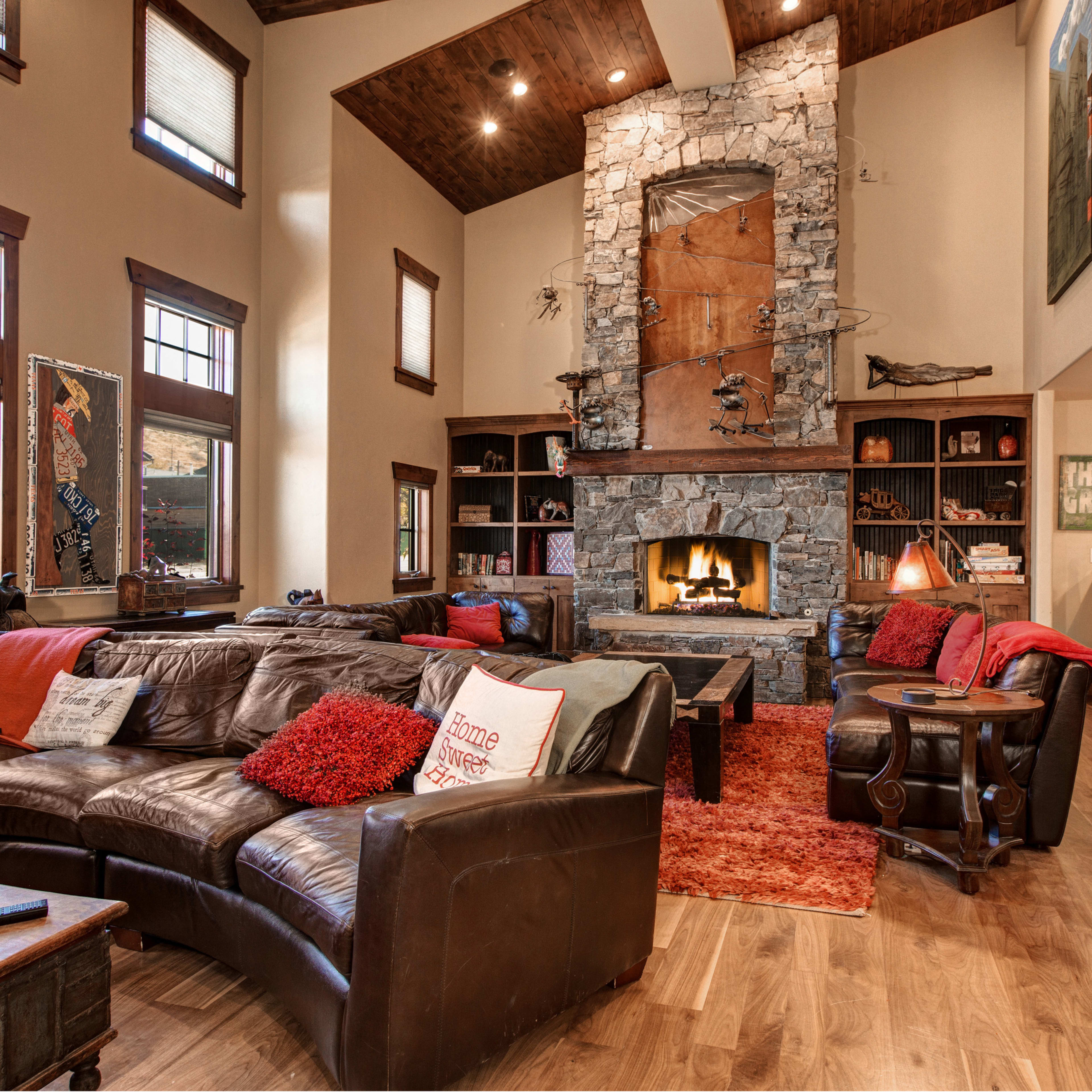 A spacious living room with a stone fireplace surrounded by a leather sectional sofa, wooden ceiling beams, and large windows.