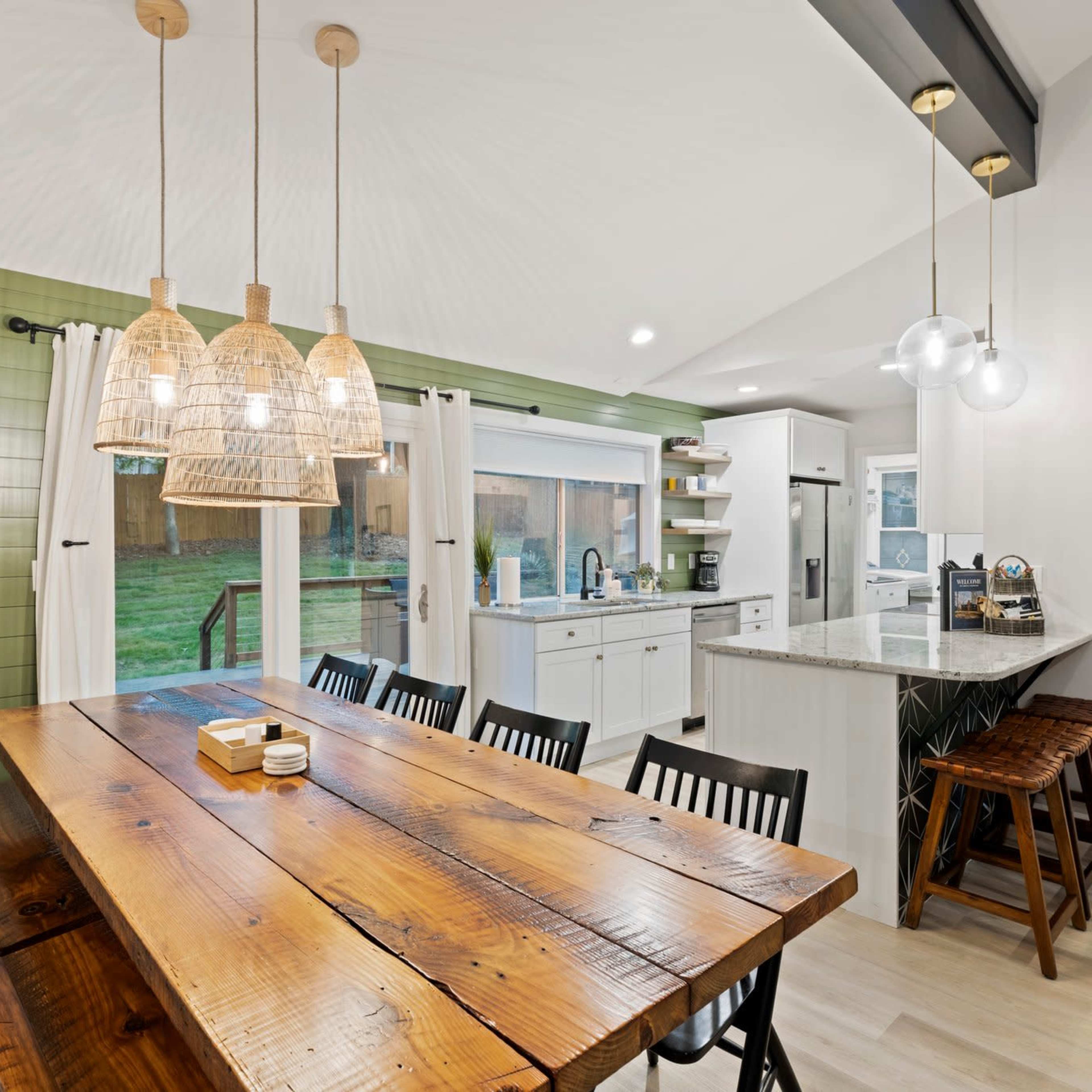 A modern kitchen features a large wooden dining table, pendant lights, and a green accent wall with a large window overlooking a backyard.