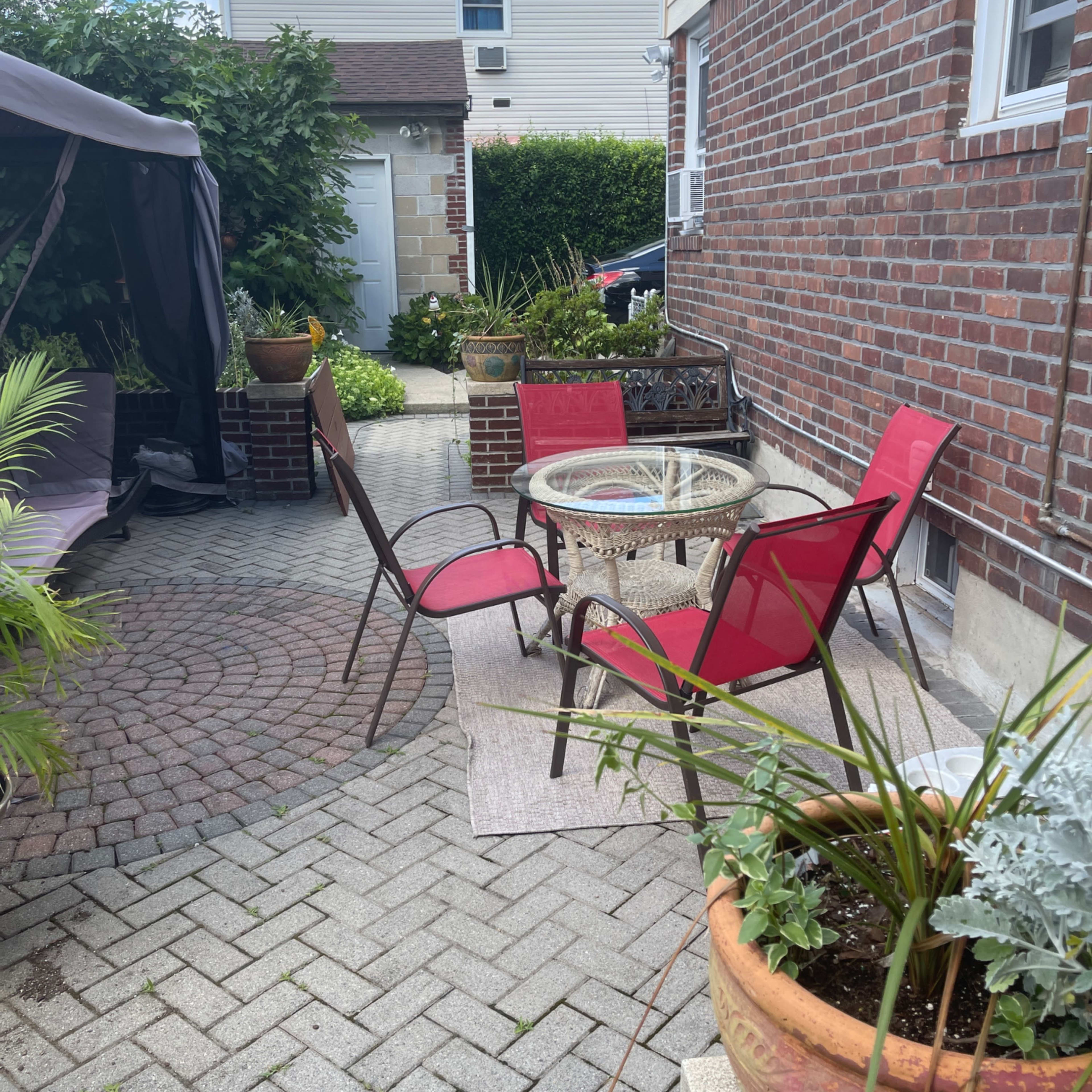 A small patio area features a round glass table surrounded by four red chairs, with brick walls and greenery in the background.