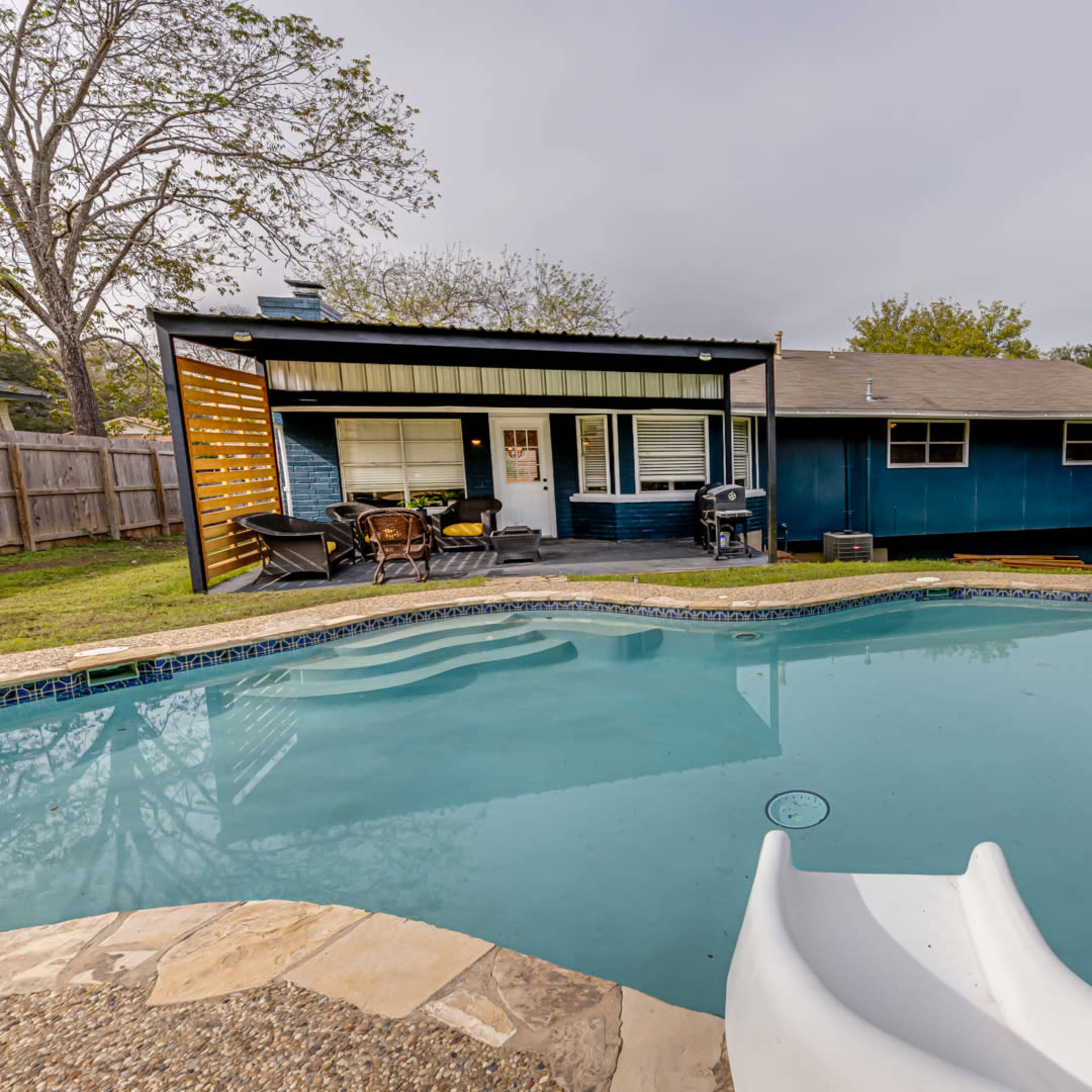 A backyard scene featuring a swimming pool with a slide, adjacent to a single-story house with a patio and yard.