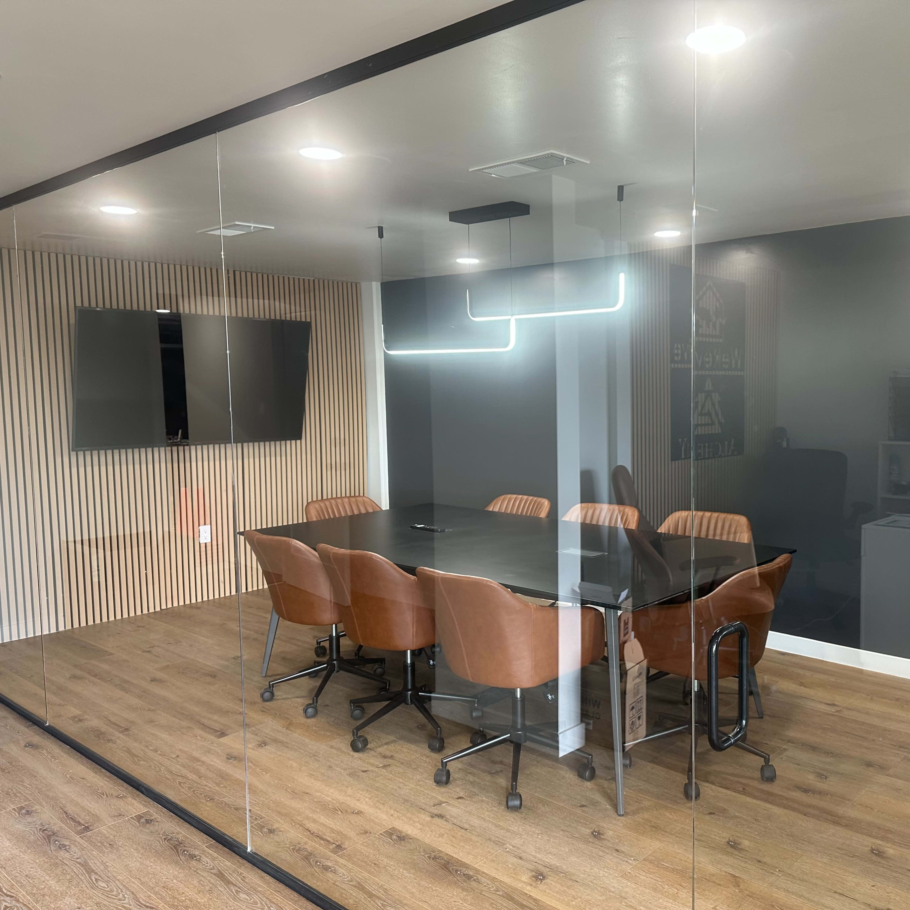 A modern conference room with a large black table surrounded by brown chairs, glass walls, and a wall-mounted TV.