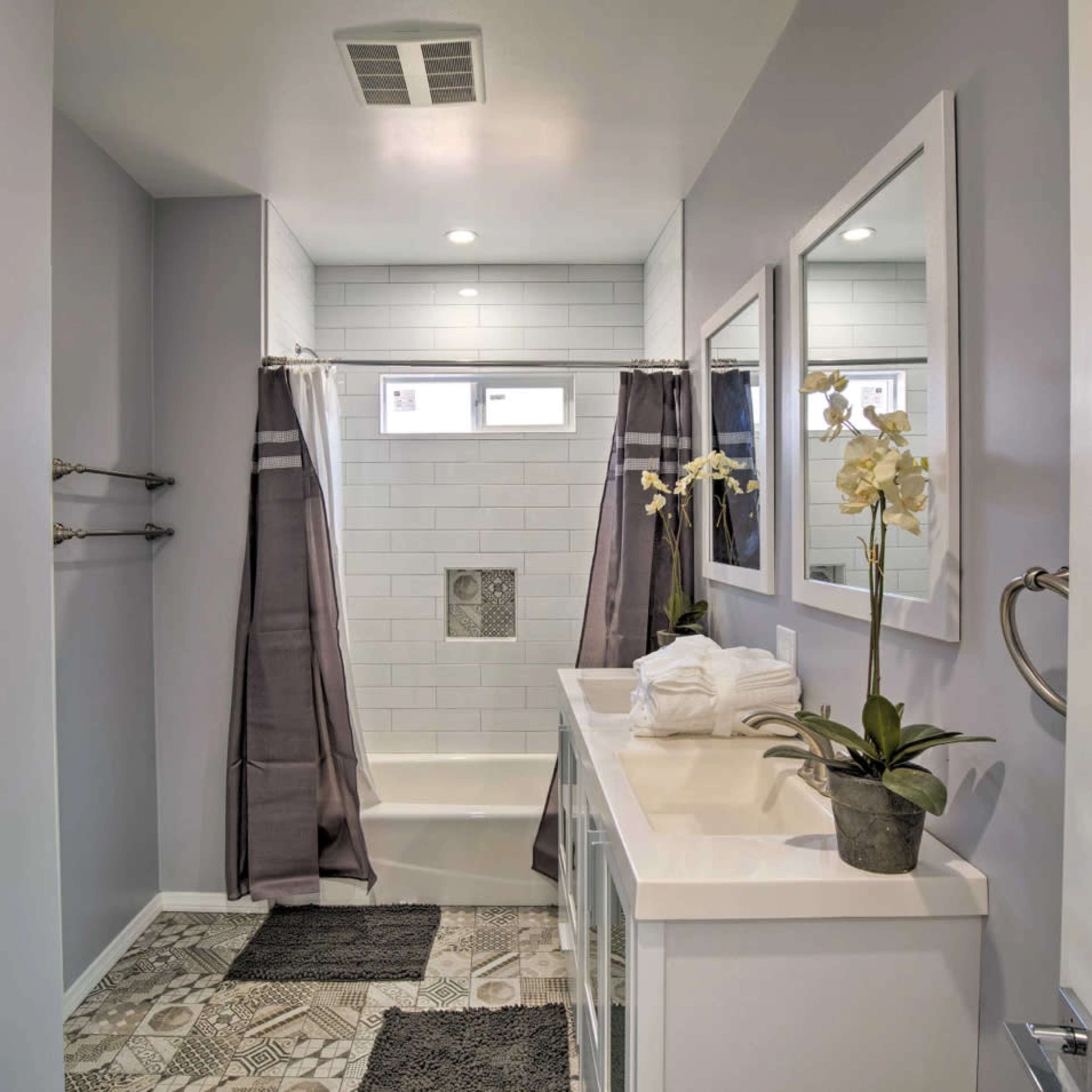 A modern bathroom featuring a bathtub with a shower, gray curtains, two mirrors, and a vanity with neatly arranged towels and a potted plant.