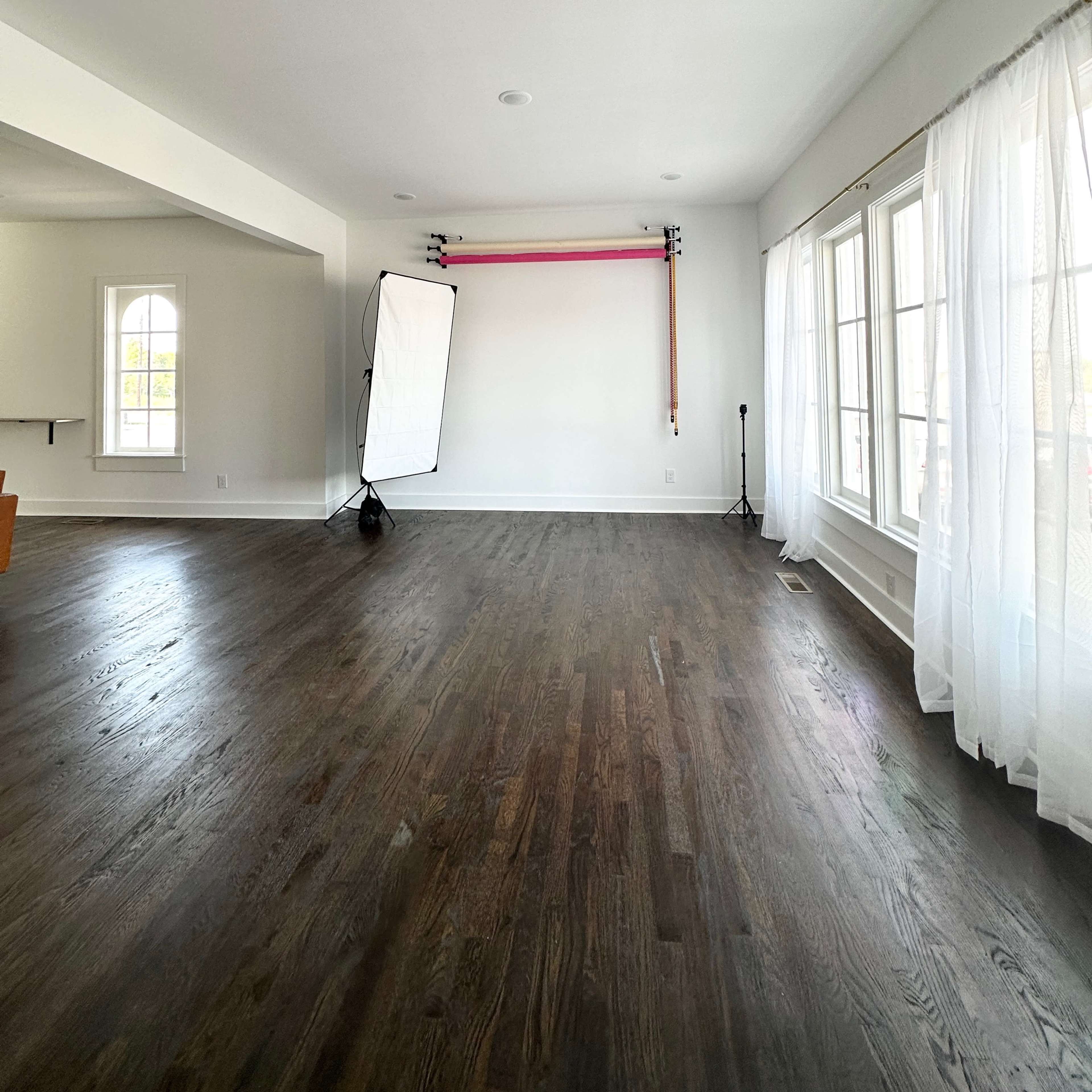 A spacious, empty room with dark hardwood floors and large windows covered by sheer white curtains, featuring a backdrop setup on one wall.
