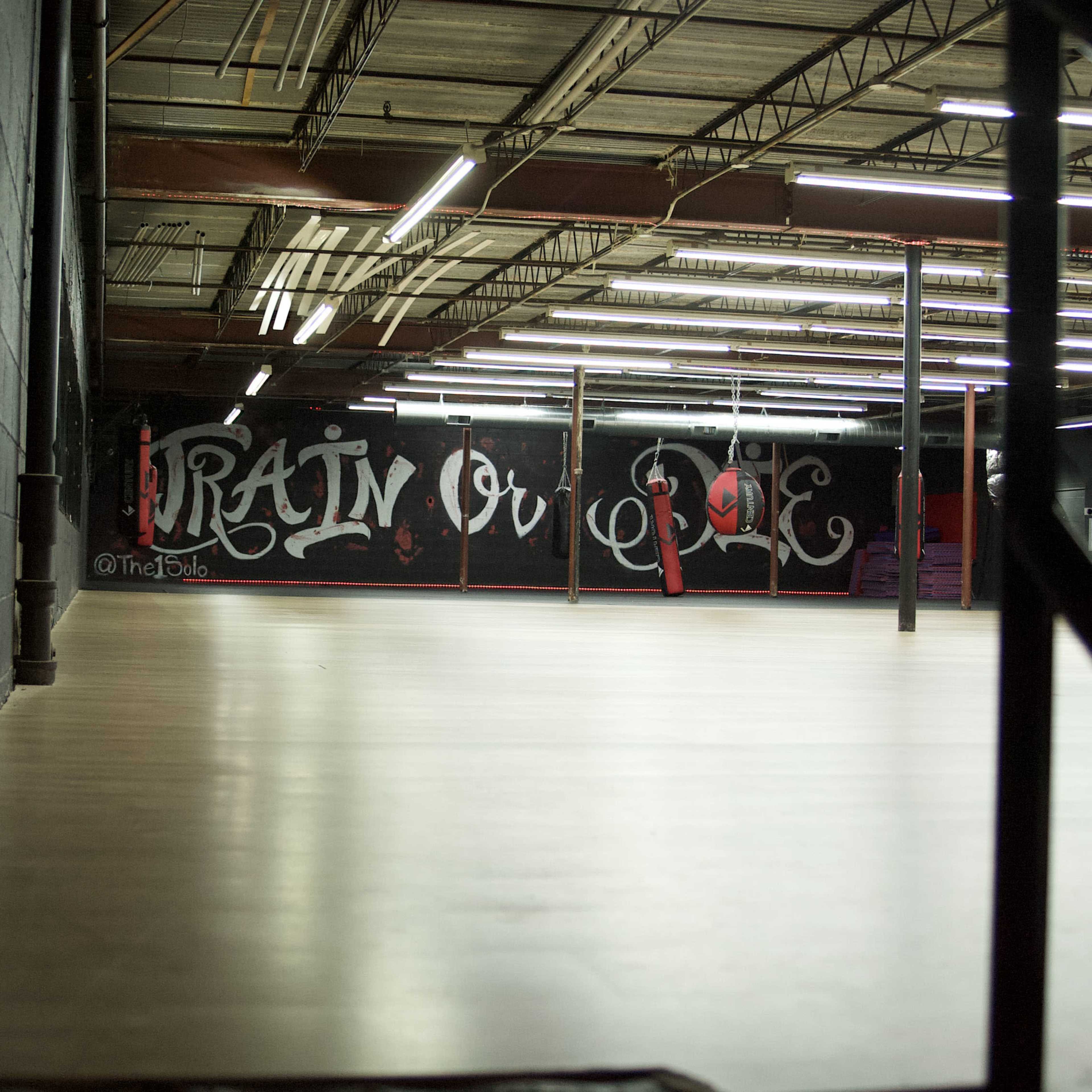 An empty indoor space with a polished wooden floor and a wall featuring graffiti that says "Rain or Die."