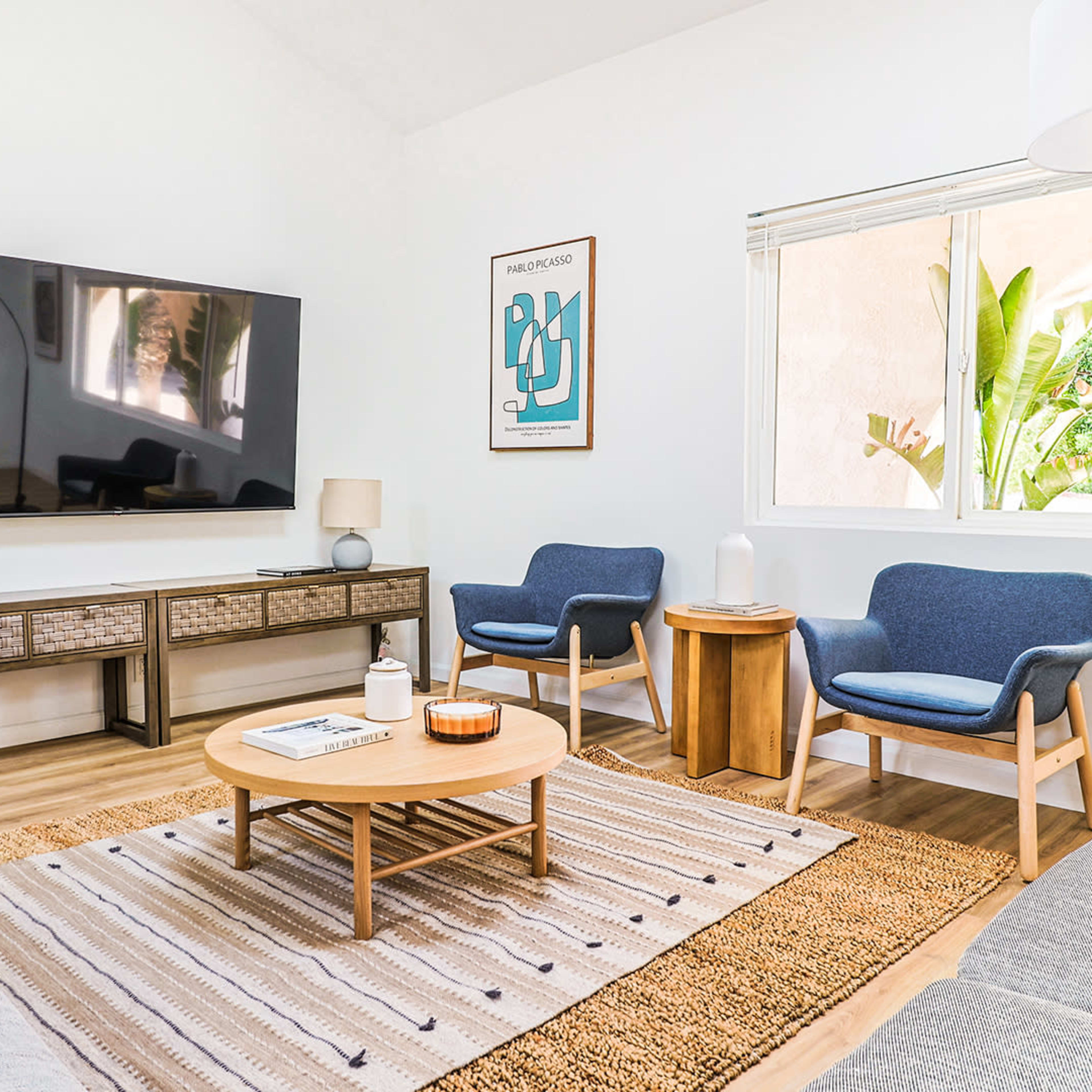 A modern living room featuring a television mounted on the wall, two blue chairs, a circular coffee table, and natural light coming through a window.