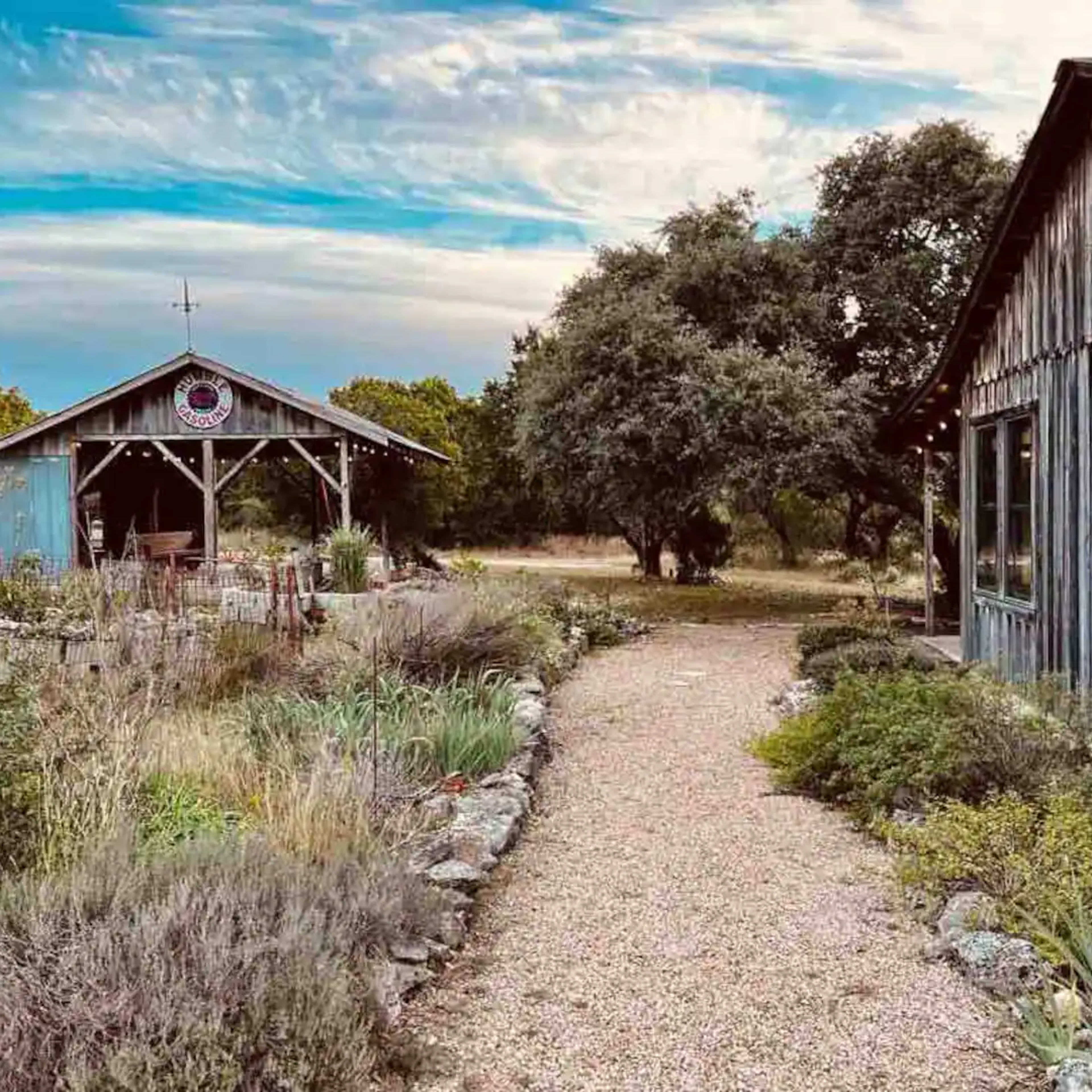 A gravel path winds between two rustic buildings amidst landscaped gardens and trees under a cloudy sky.