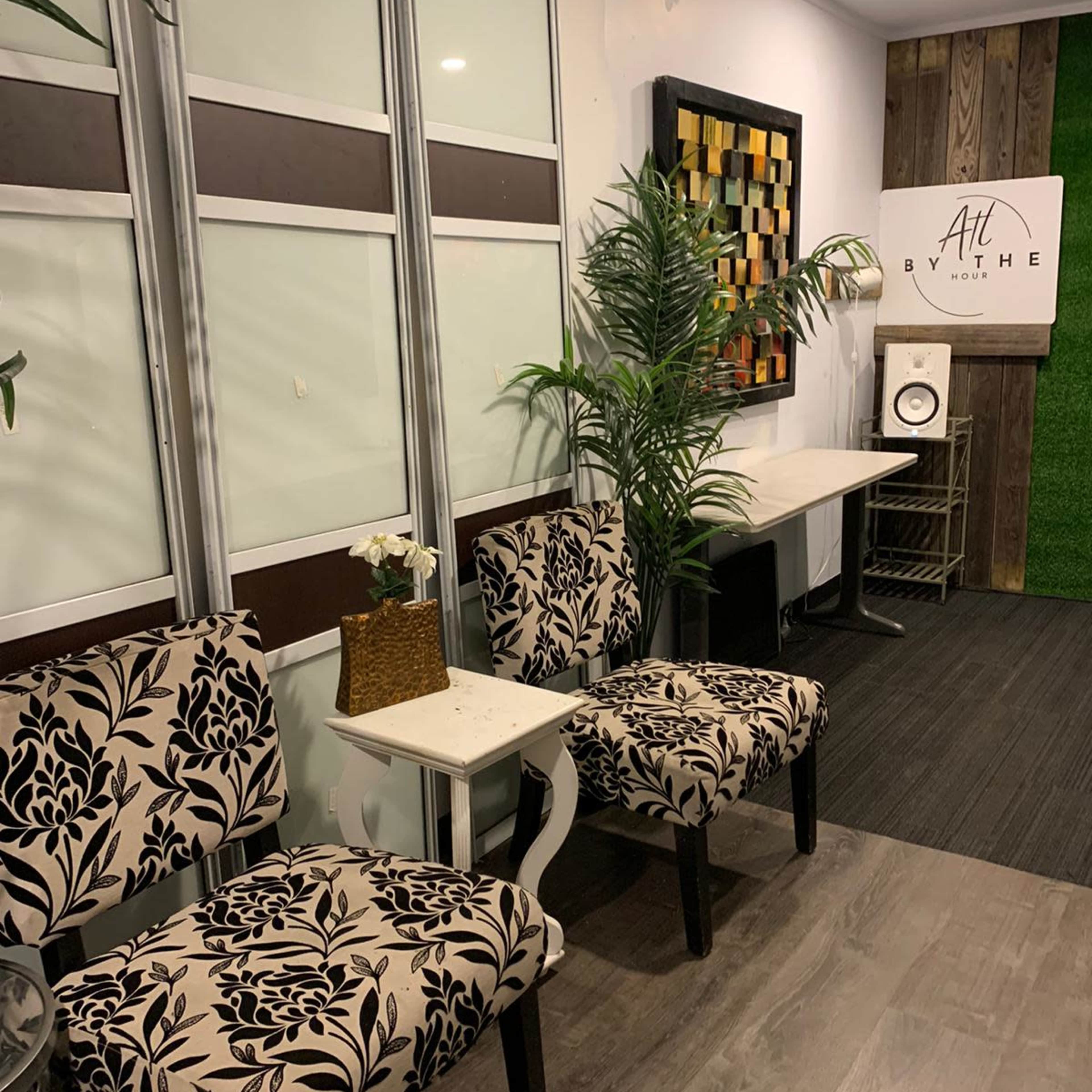 A small seating area featuring two patterned chairs, a white side table, and decorative plants against a backdrop of wooden and white walls.