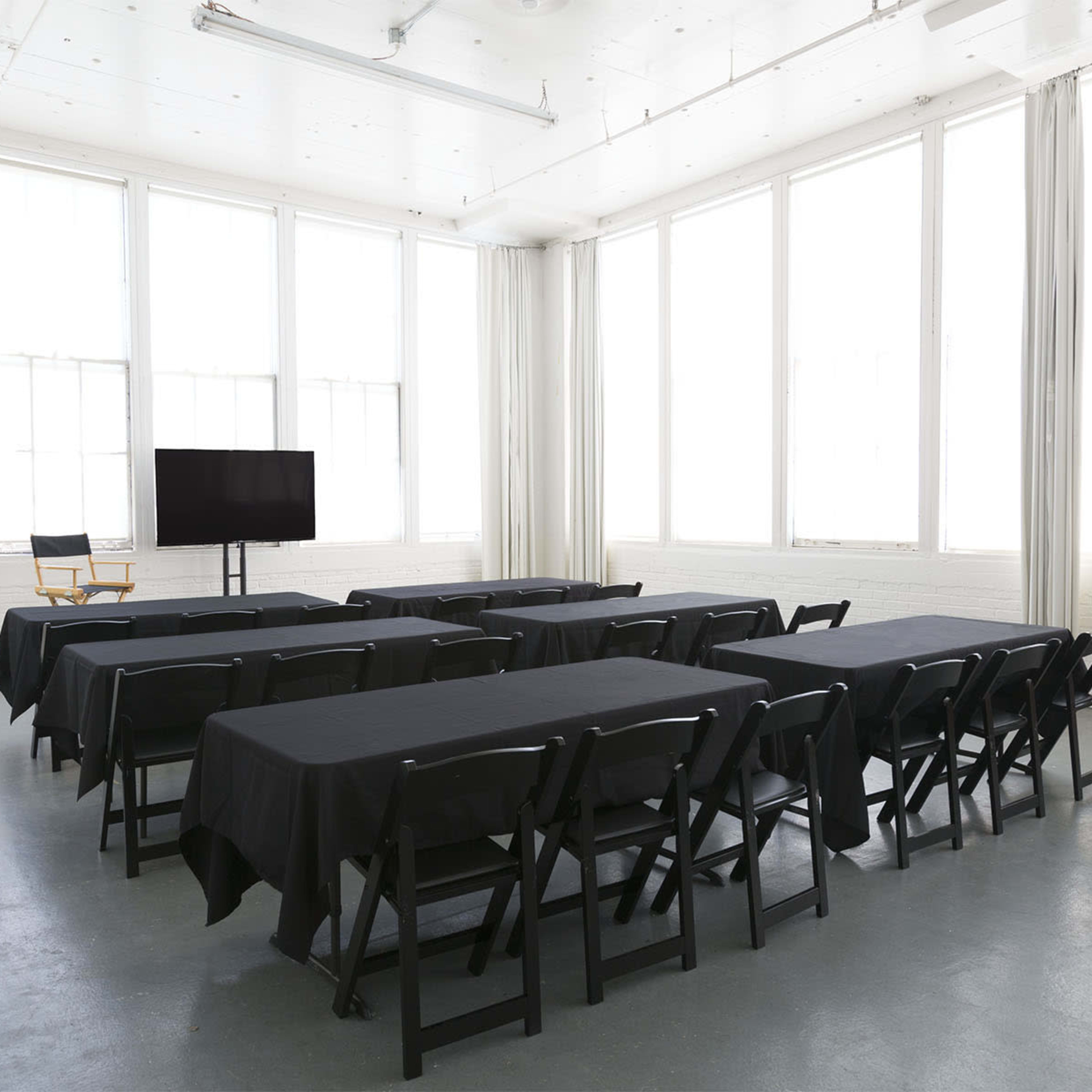 A bright, minimalist meeting room set up with several rows of black tables and chairs, facing a blank screen on the front wall.