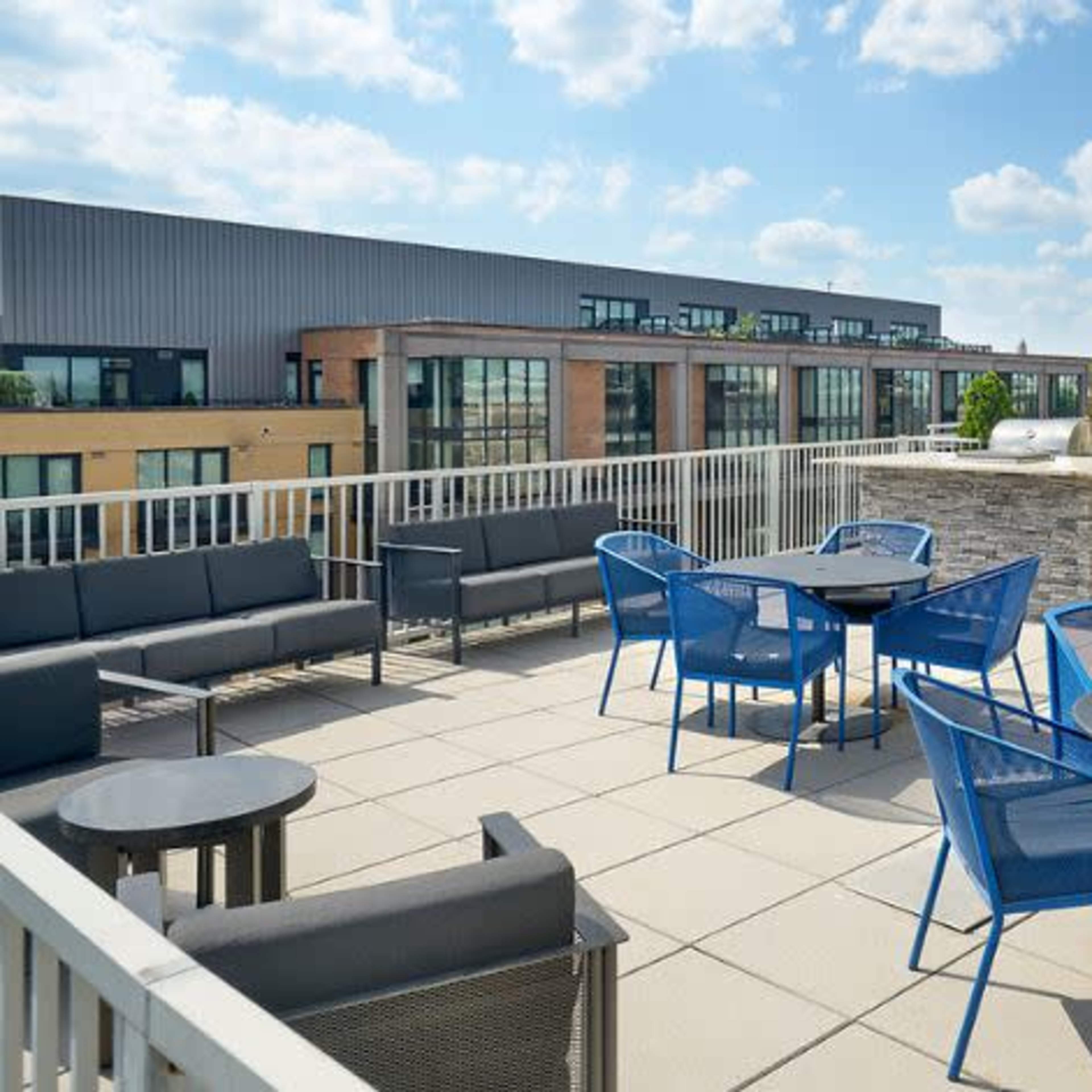 A rooftop terrace featuring several blue patio chairs and tables, along with gray couches, surrounded by buildings under a partly cloudy sky.